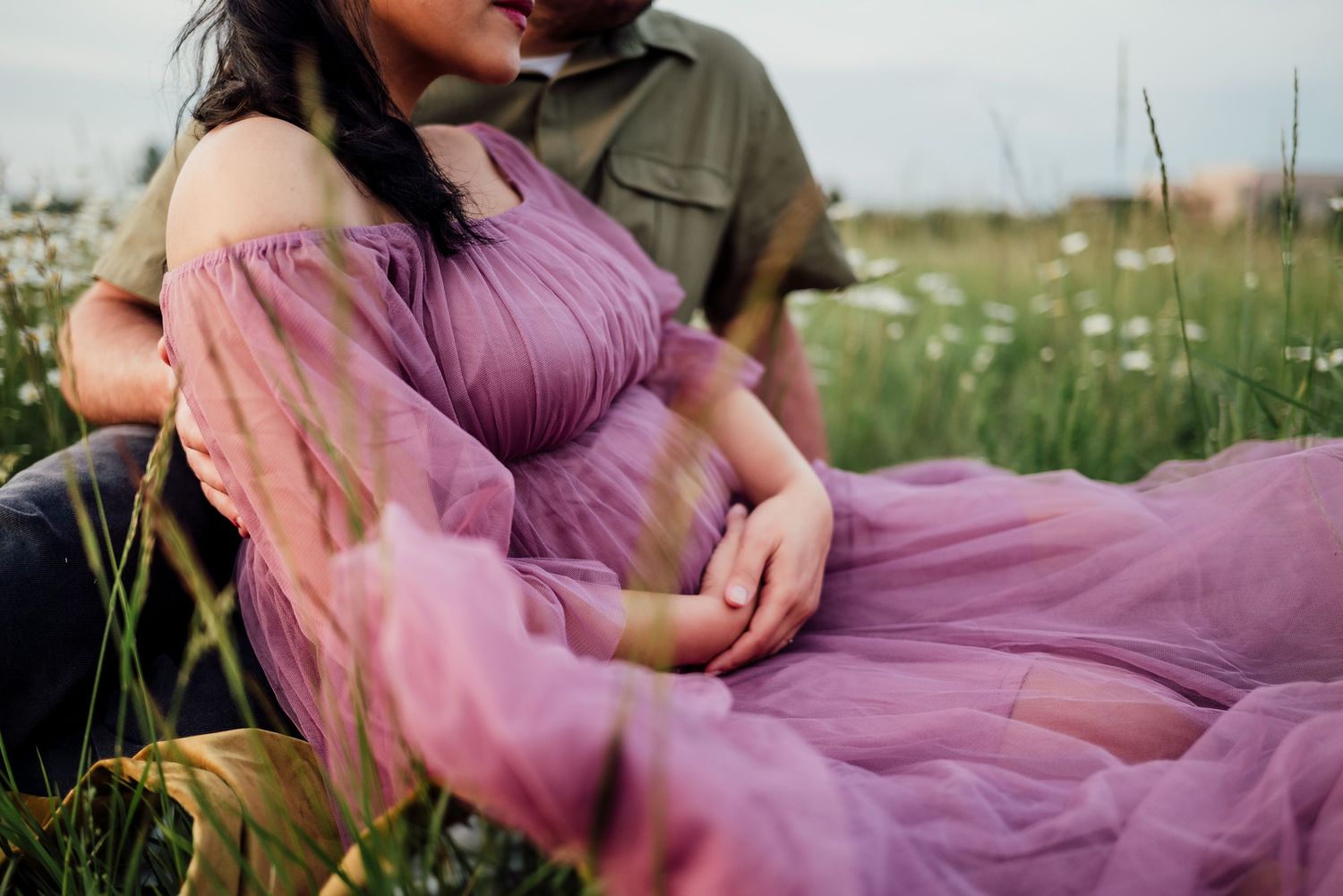 Pregnant woman in purple dress sitting in wildflower field with partner during maternity photo session.