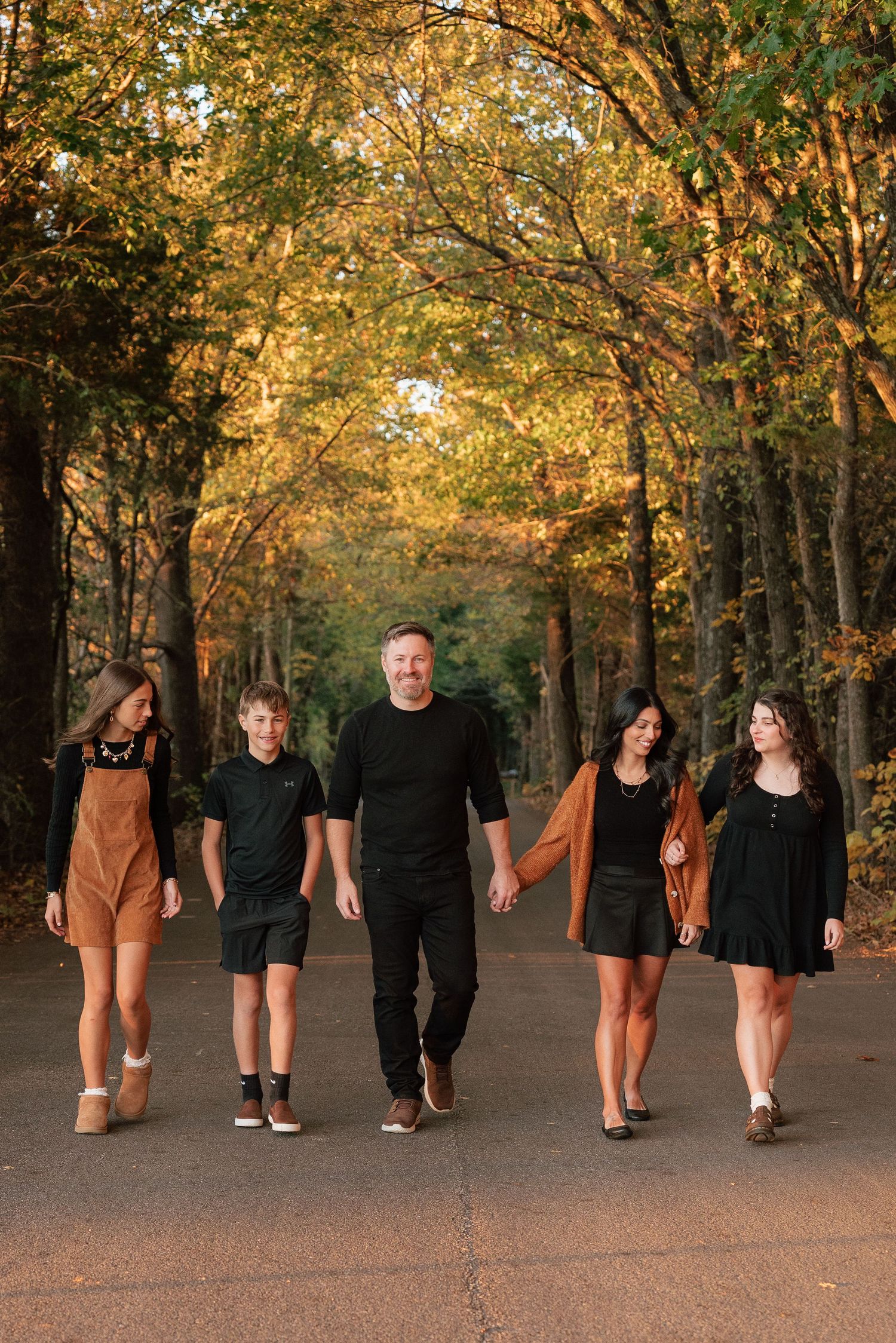 Family of five walking together on a tree-lined path surrounded by colorful autumn foliage.