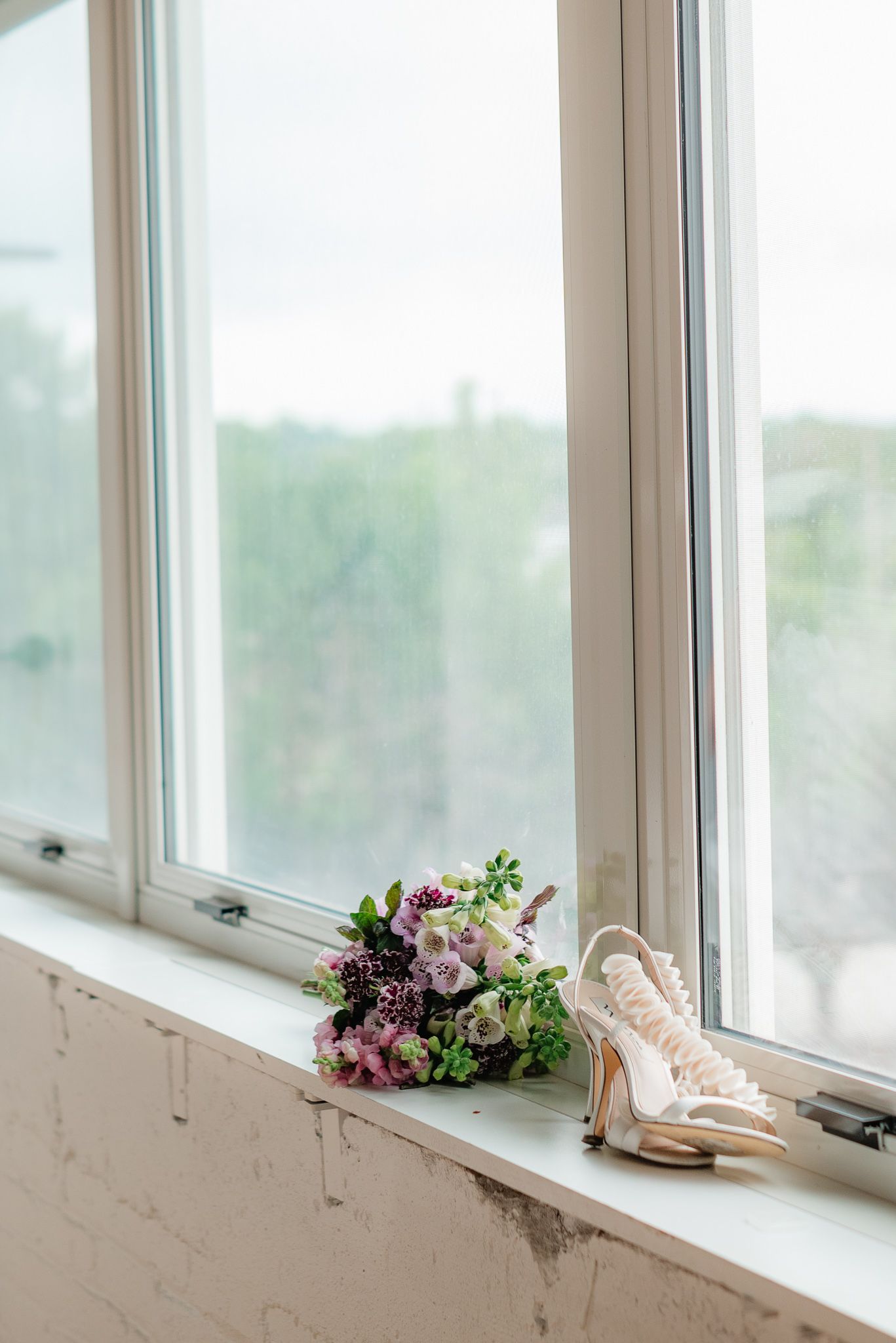 Bridal bouquet with purple and pink flowers beside lace heels on a white windowsill with garden view.