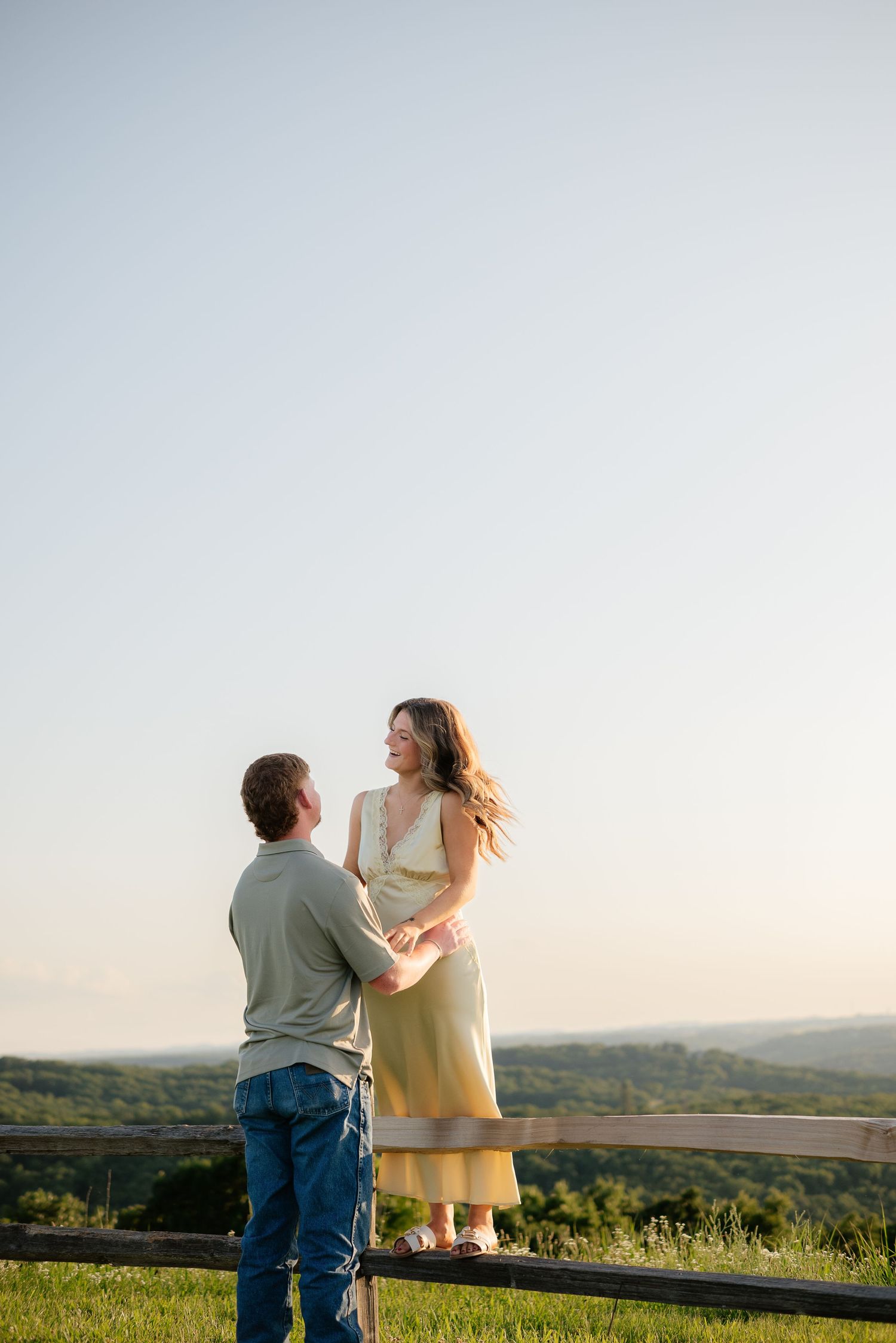 Couple holding hands on a grassy hilltop at golden hour, smiling at each other with scenic rolling hills in the background.