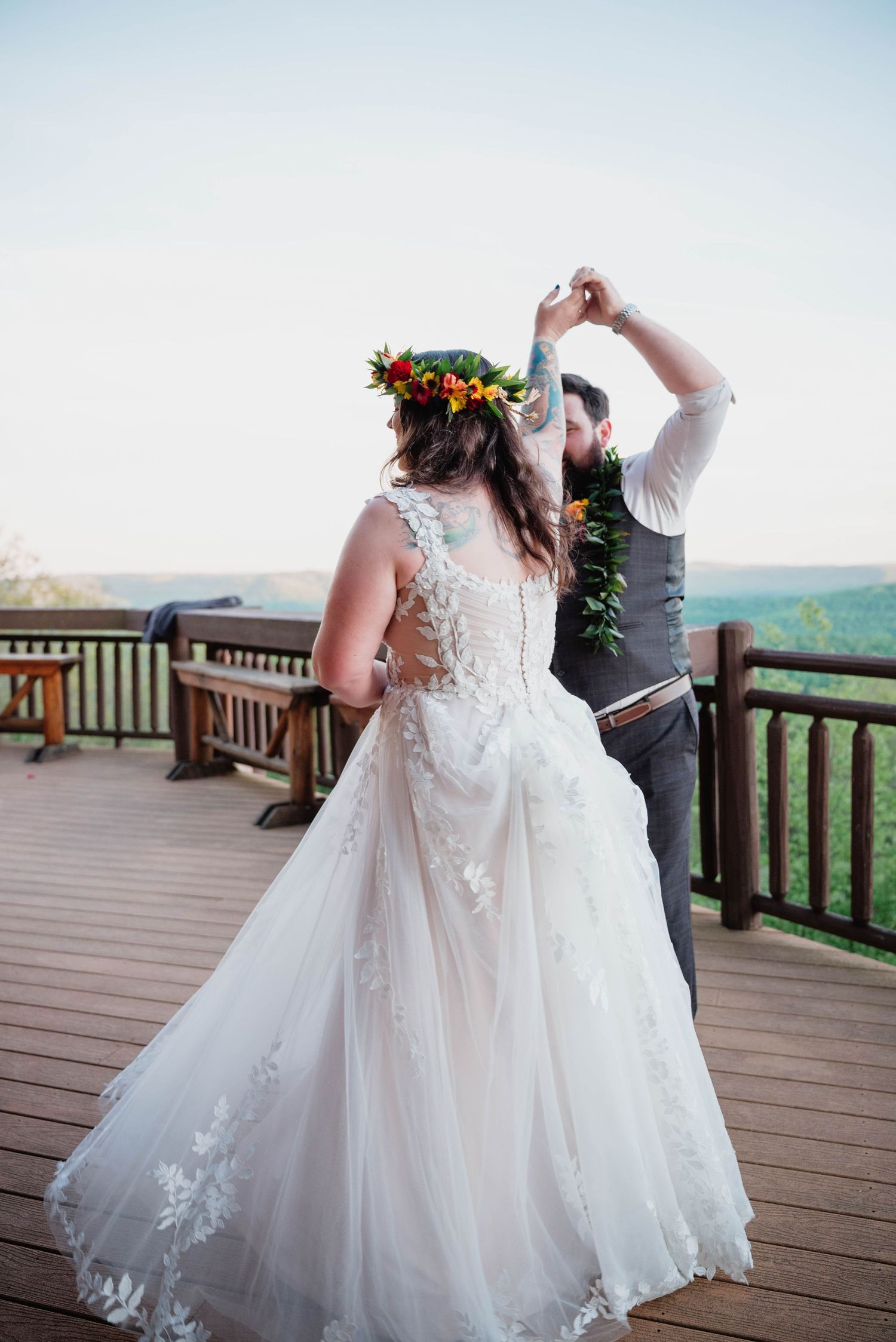 Bride in lace gown and floral crown dances with groom in gray suit on wooden deck overlooking scenic landscape.