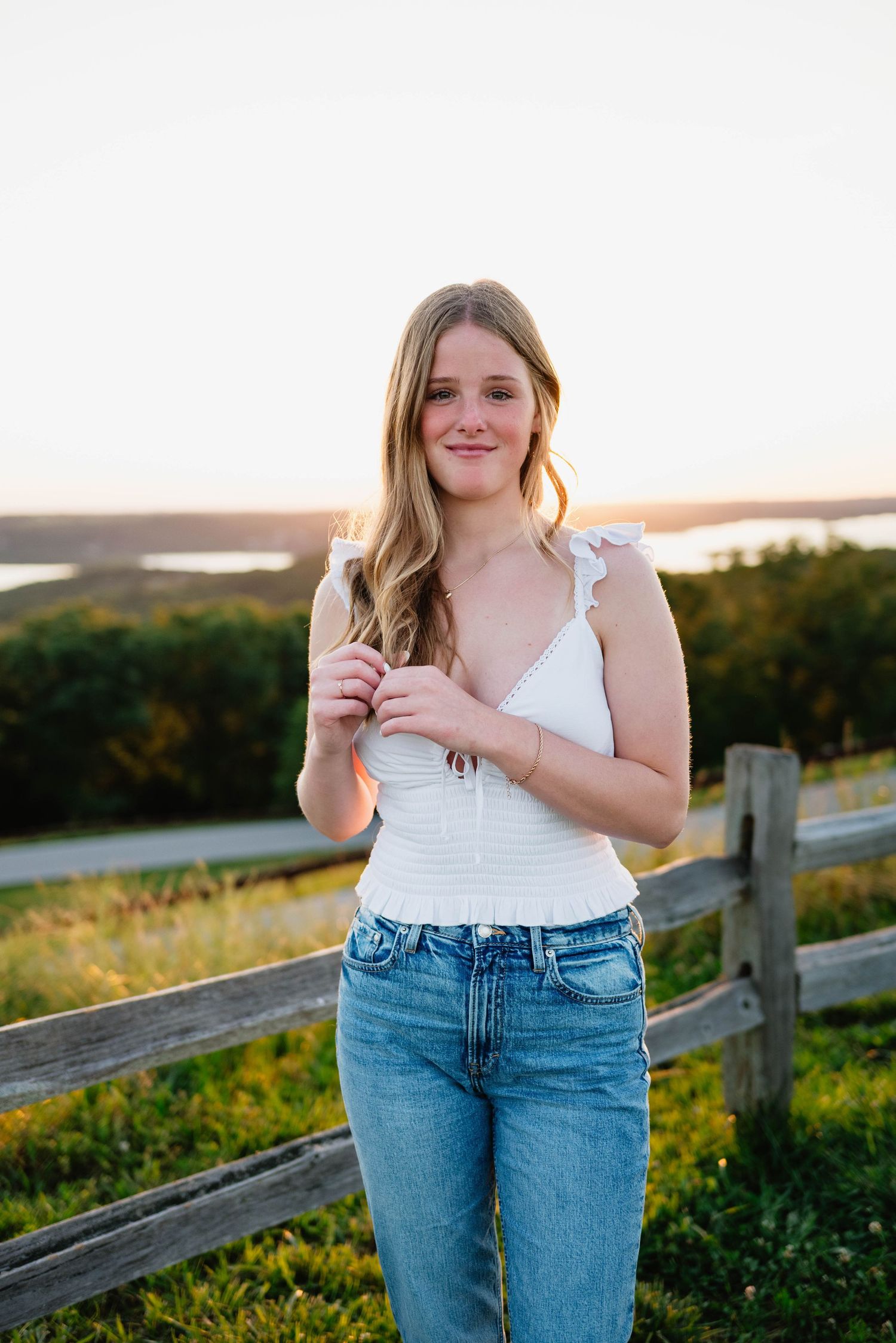 Young woman in white top and jeans poses by wooden fence at golden hour with scenic overlook behind her.