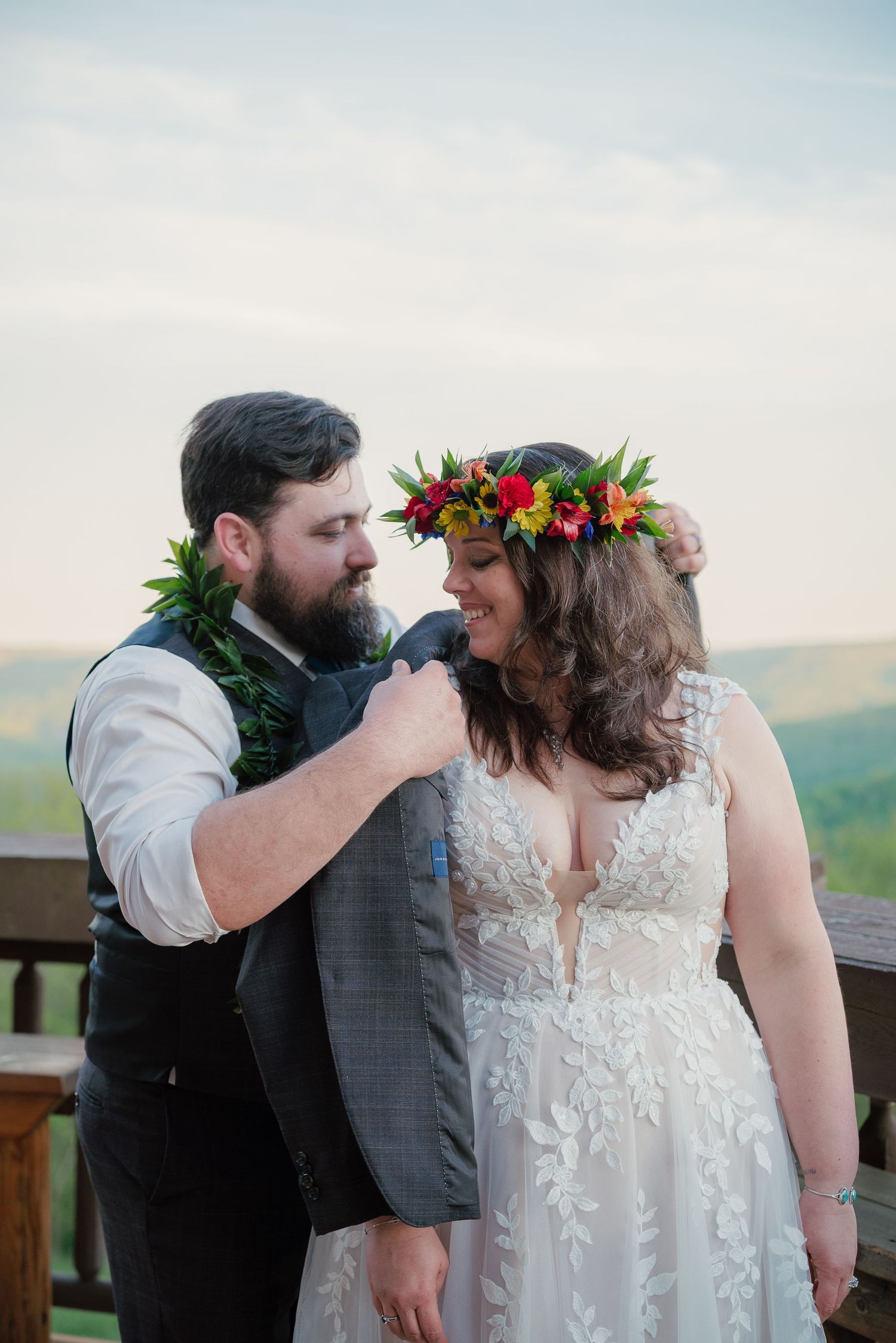 A bearded groom in a gray vest and a bride wearing a vibrant floral crown share a tender moment outdoors.