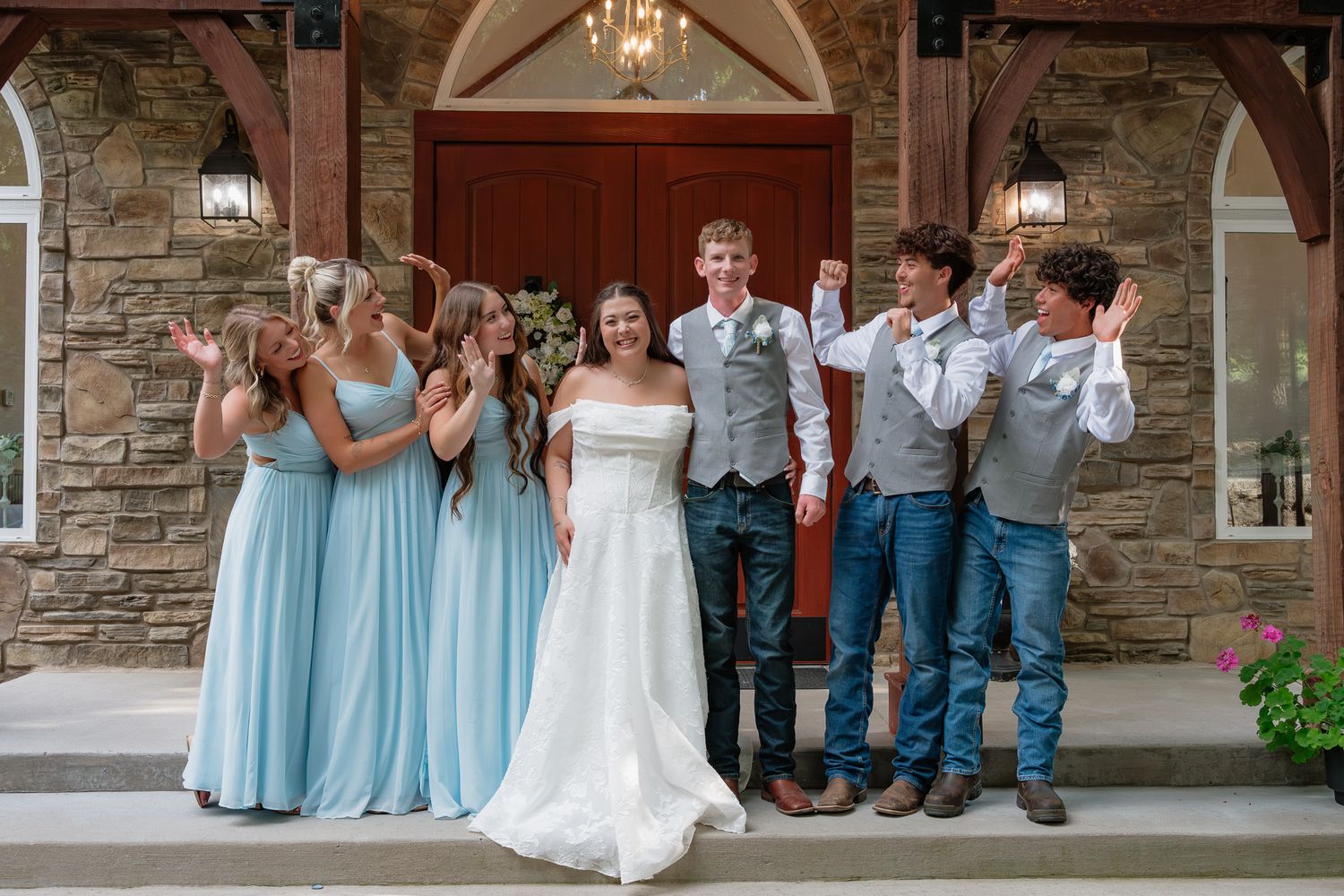 Bride and groom celebrate with bridesmaids in blue gowns and groomsmen in jeans outside a stone chapel.