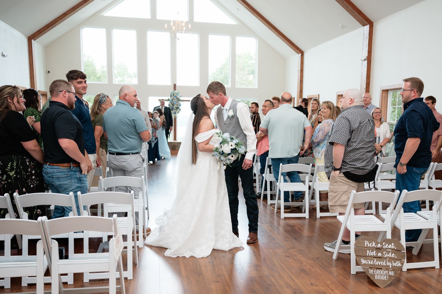Bride and groom share a kiss at the altar during an indoor wedding ceremony with white chairs and guests standing in a bright venue.
