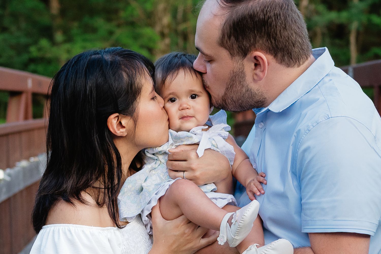 Parents kissing their baby on the cheeks on a wooden bridge outdoors.
