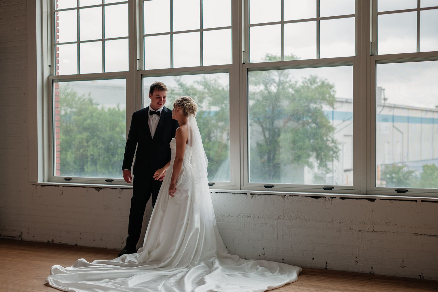Bride and groom share an intimate moment standing by large industrial windows in a bright wedding venue.