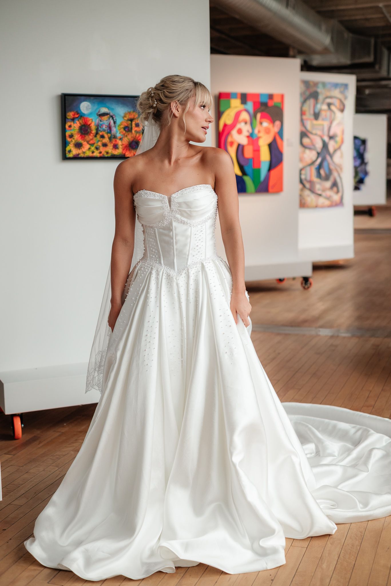 Bride in strapless white ball gown with corset bodice and train poses in art gallery with colorful paintings.