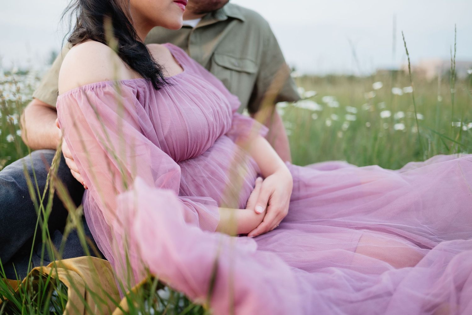 Pregnant woman in purple dress sitting in flower field with partner during maternity photoshoot.