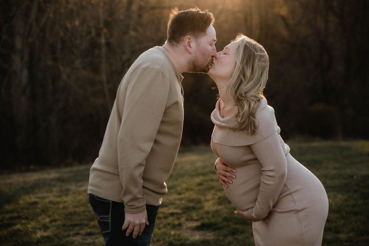 Couple sharing a tender kiss outdoors at golden hour, woman in beige maternity dress showing baby bump.