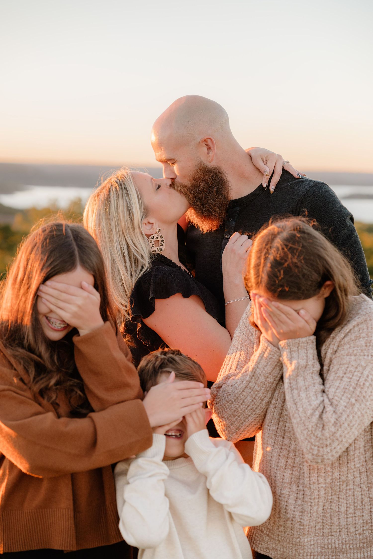 A couple shares a kiss at golden hour while three children playfully cover their eyes outdoors.