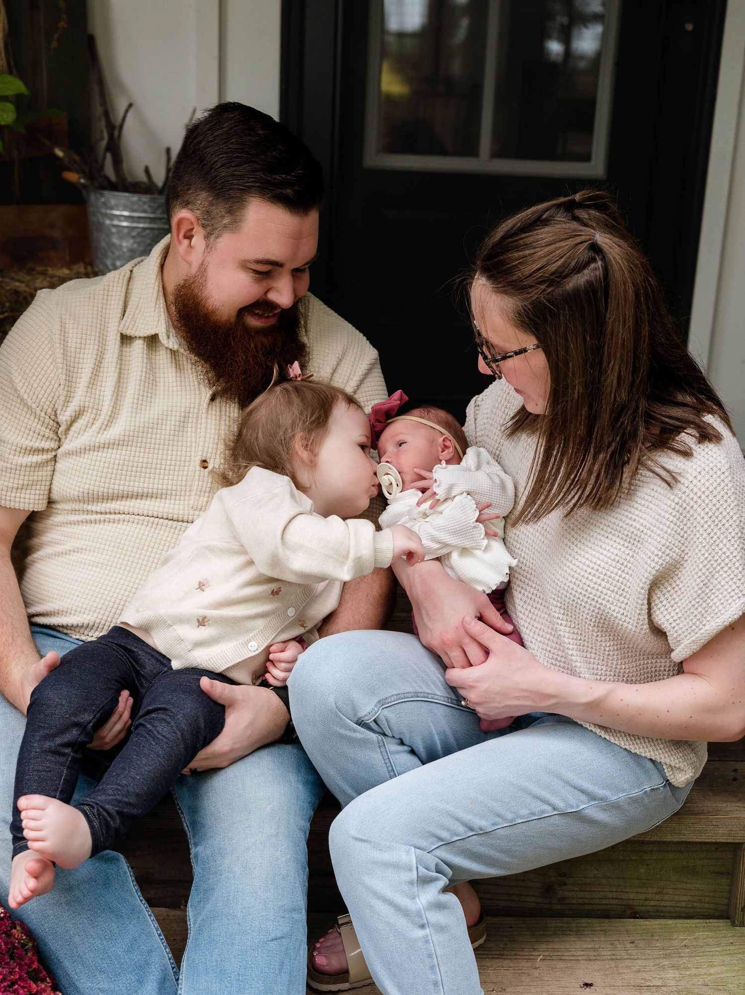 A bearded father and mother sit on porch steps with toddler and newborn baby, sharing a tender family moment.