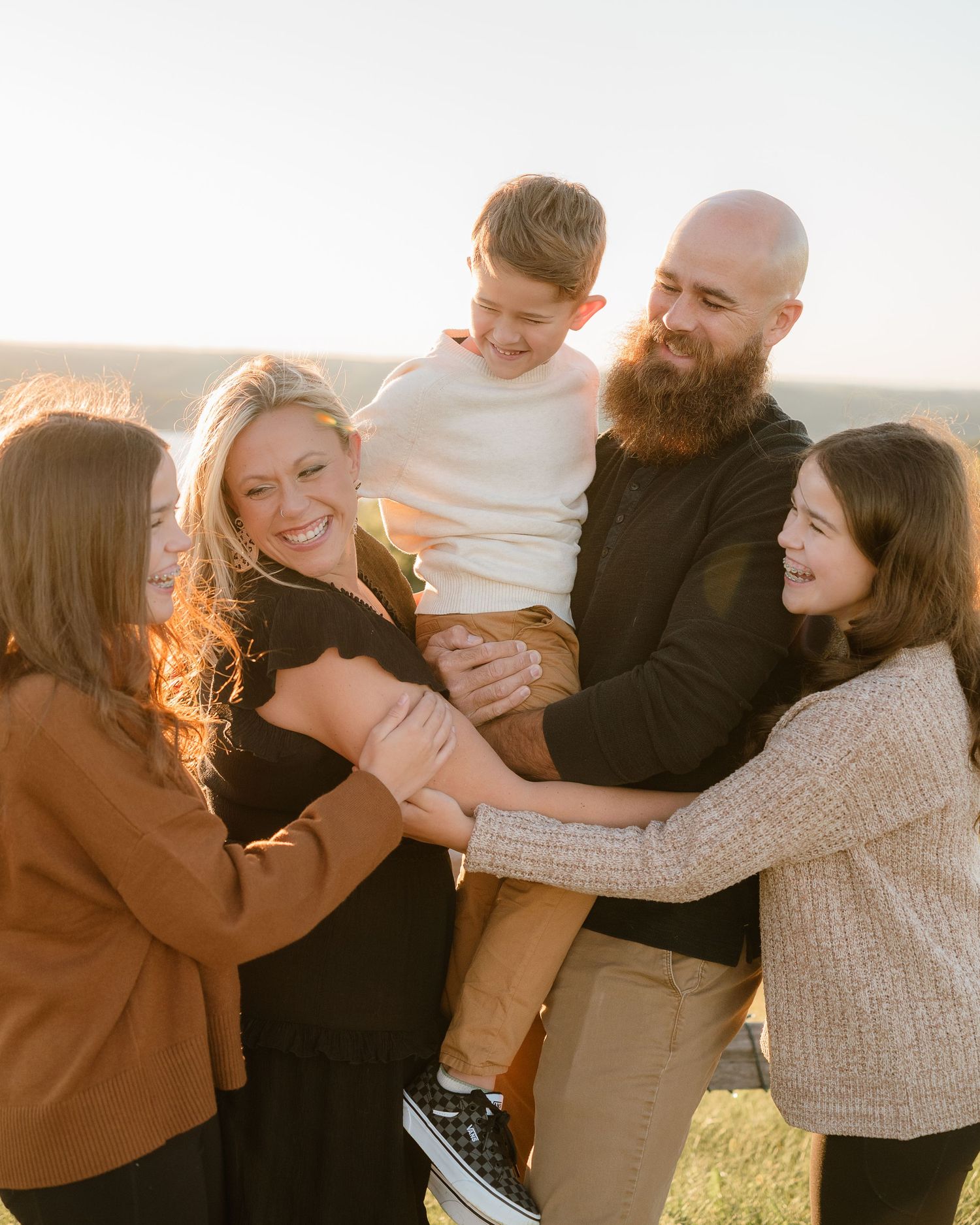 A joyful family of five laughing together outdoors at golden hour, with a bearded dad holding a young boy.