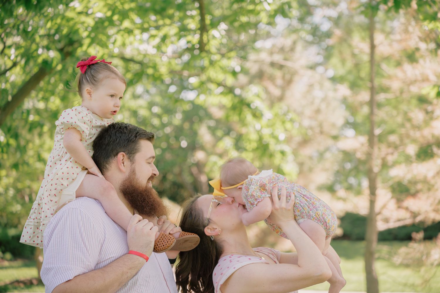 Happy family with bearded dad, mom, and two young daughters enjoying a sunny day outdoors in a lush green park.