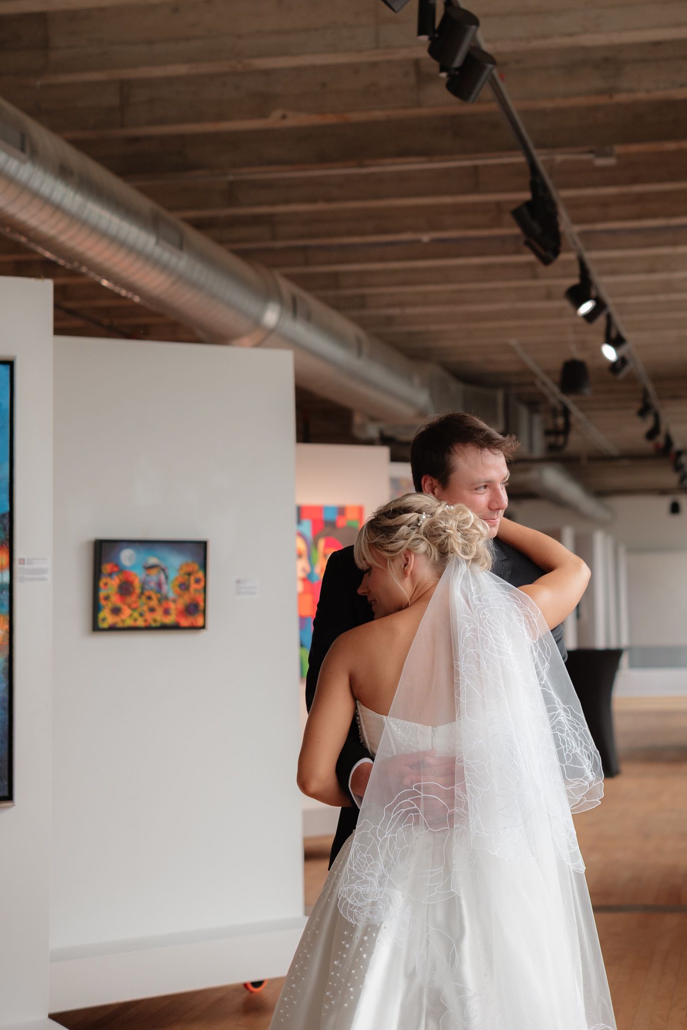 Bride and groom share a tender embrace in an art gallery wedding venue with colorful paintings and industrial ceiling.