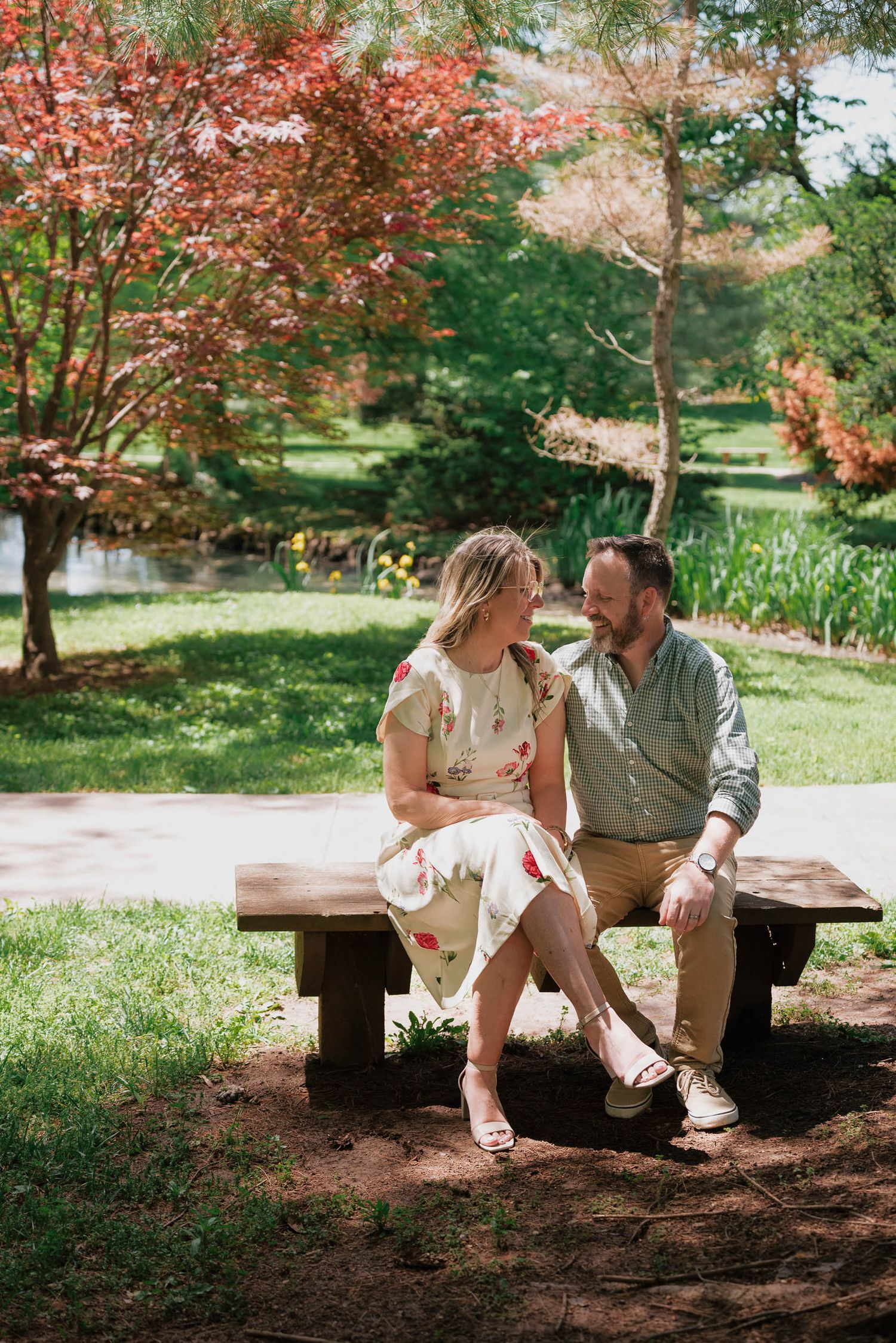 Couple sitting on a wooden bench in a lush garden park, surrounded by colorful autumn trees near a pond.