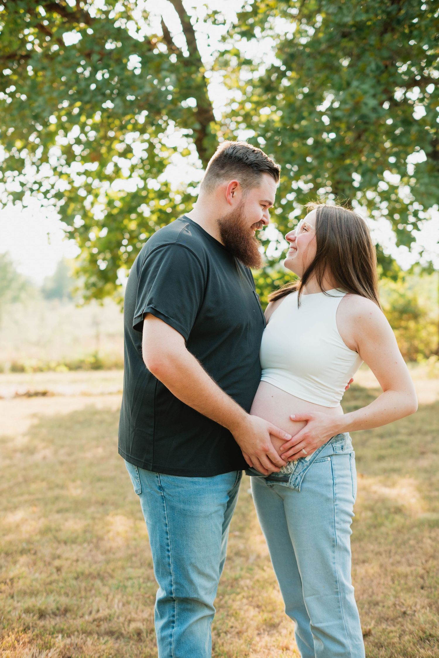 A couple smiles at each other outdoors, cradling a pregnant belly, surrounded by lush green trees in warm sunlight.