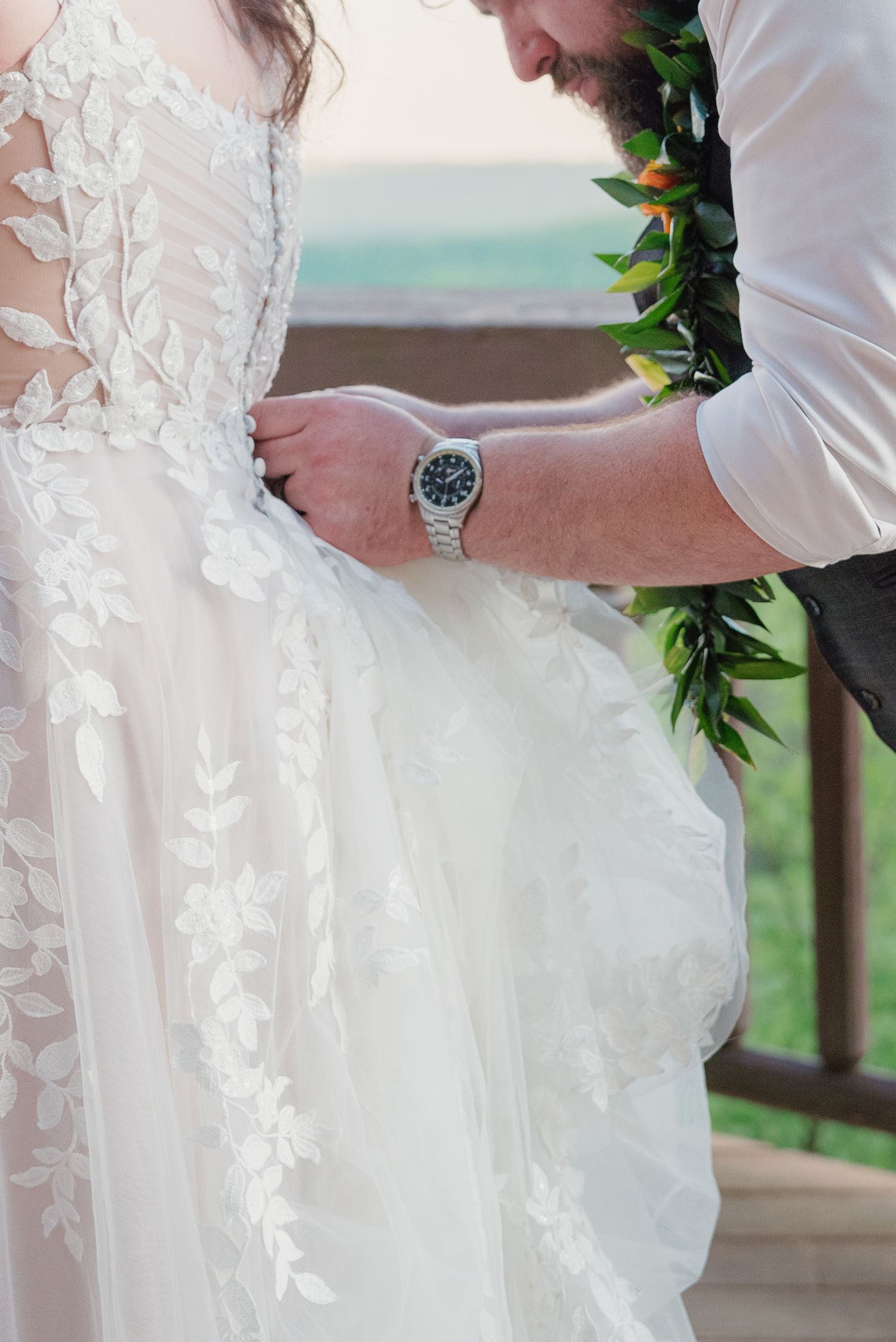 Groom in floral lei fastening buttons on bride's white lace wedding dress during outdoor Hawaii ceremony.