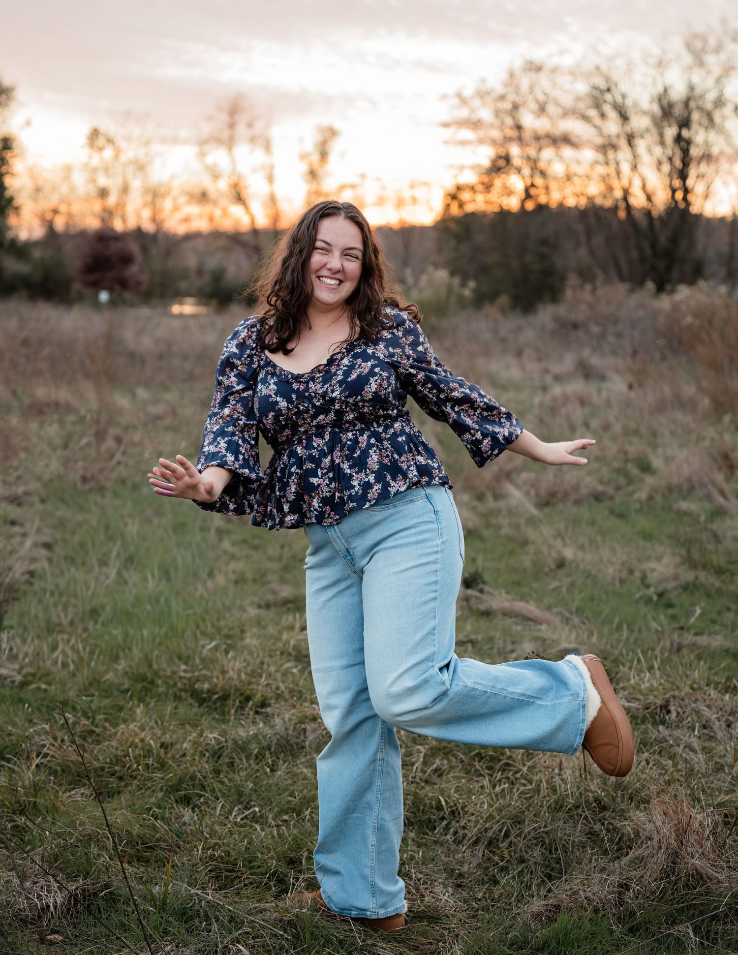 A joyful woman in floral top and wide-leg jeans poses playfully in a field at golden hour sunset.
