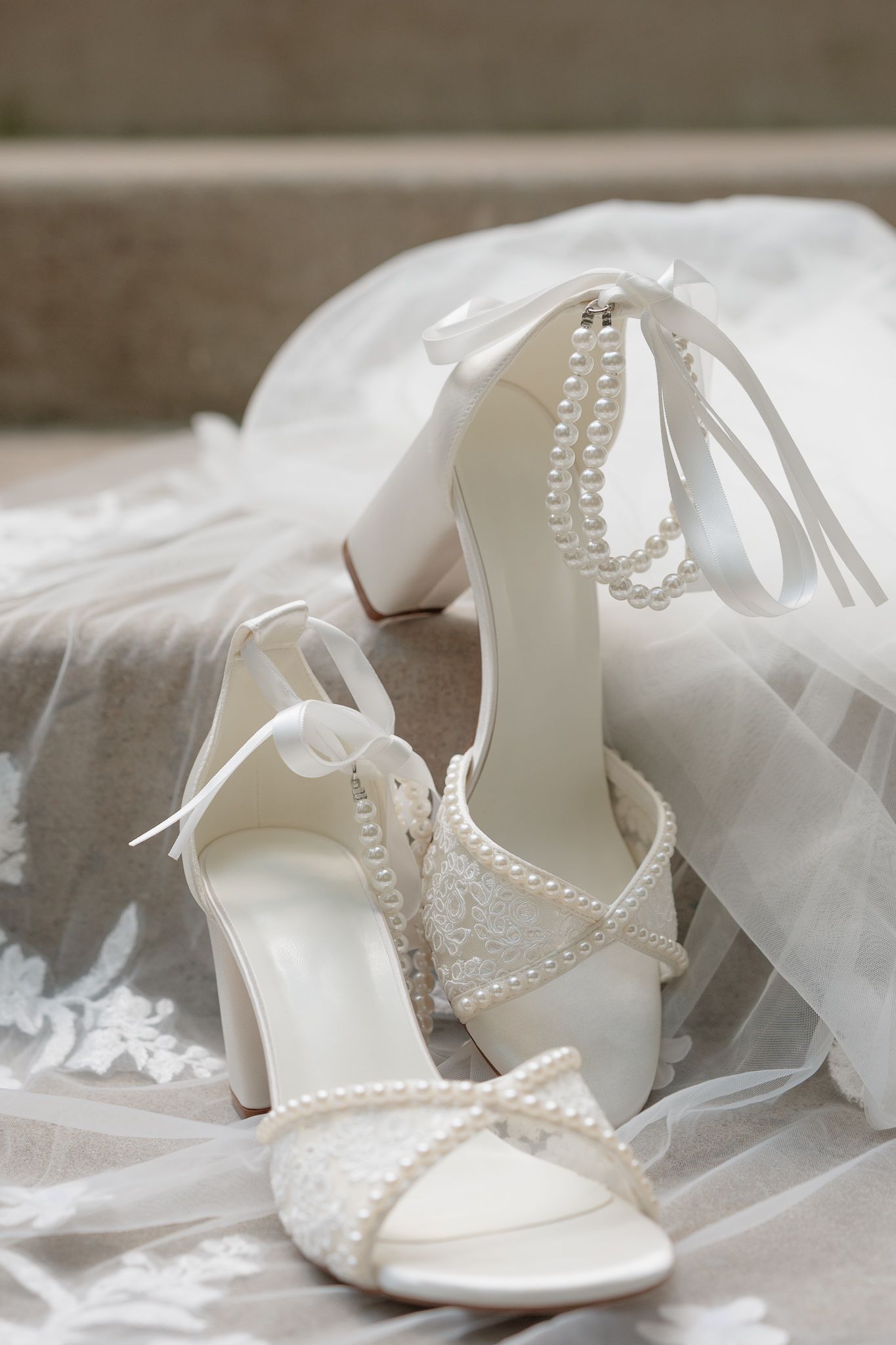 Elegant white bridal heels with pearl embellishments and satin ribbon ankle ties resting on a lace wedding veil.