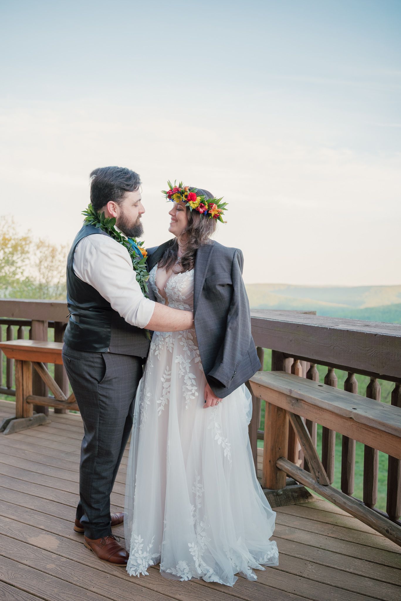 Bride in floral crown and groom with lei embrace on wooden deck with scenic mountain views on their wedding day.