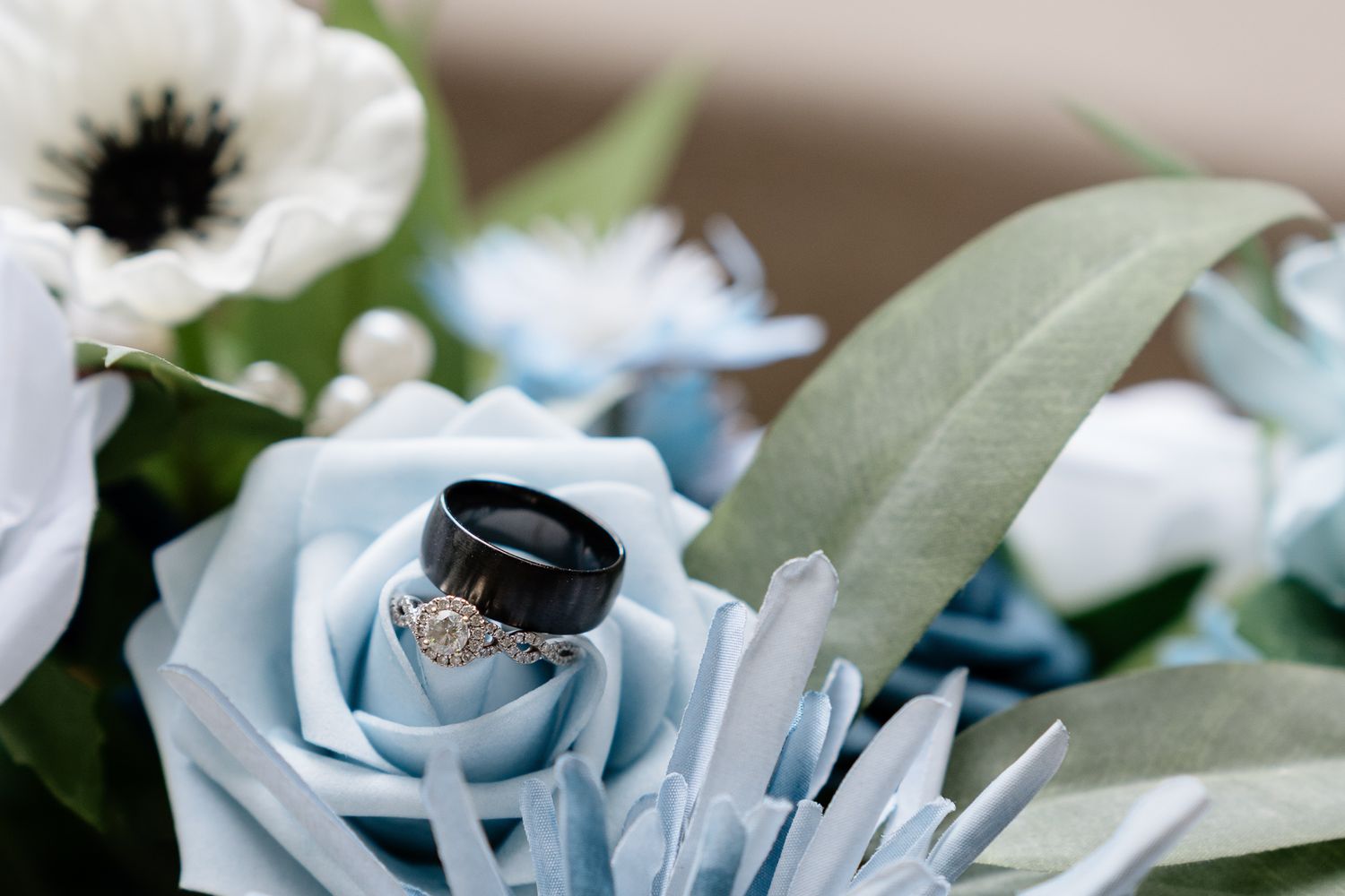 Black wedding band and diamond ring resting on light blue roses in a bridal bouquet with white anemones.