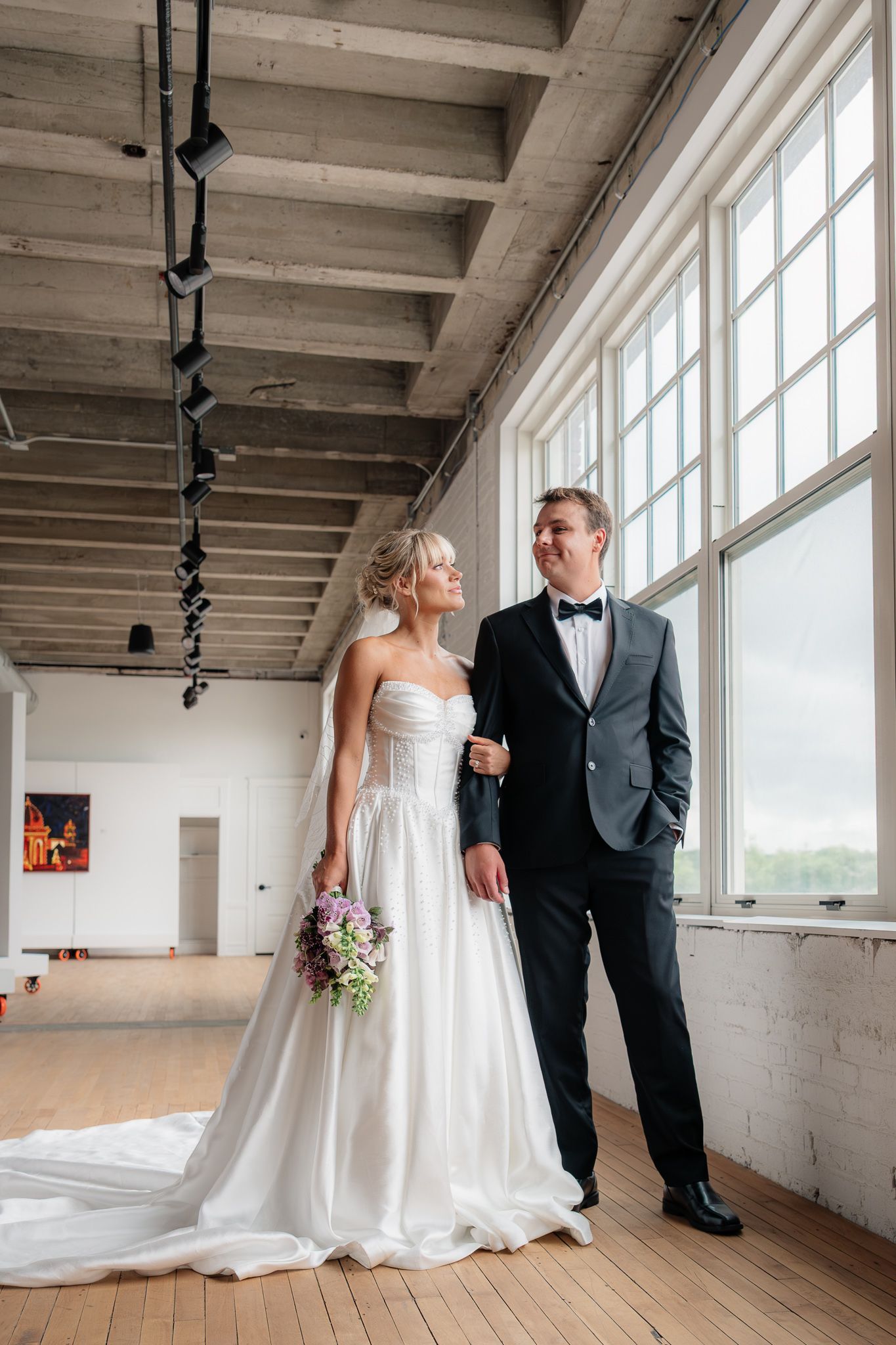 Bride in white strapless gown holding bouquet stands with groom in black tuxedo by large windows in elegant venue.