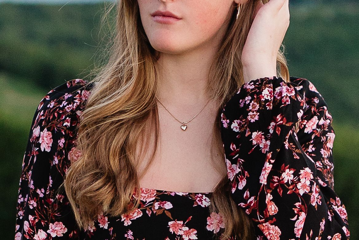 Young woman in black floral dress wearing a delicate heart necklace, posing outdoors with lush green hills behind her.