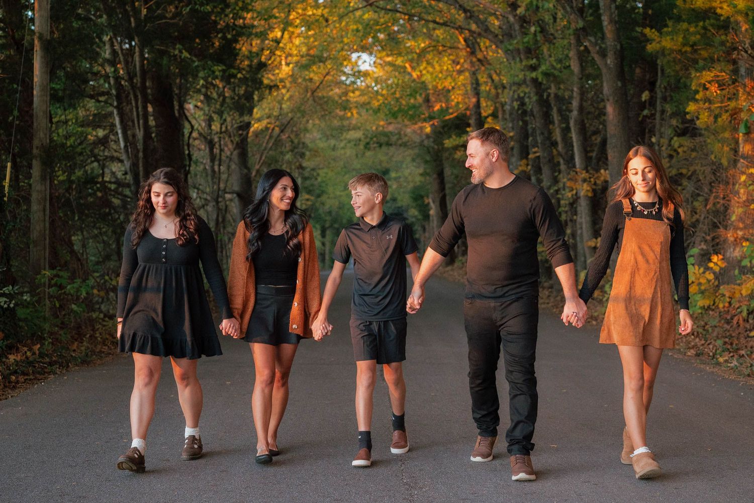 Family of five walking hand-in-hand down a tree-lined road surrounded by colorful autumn foliage.