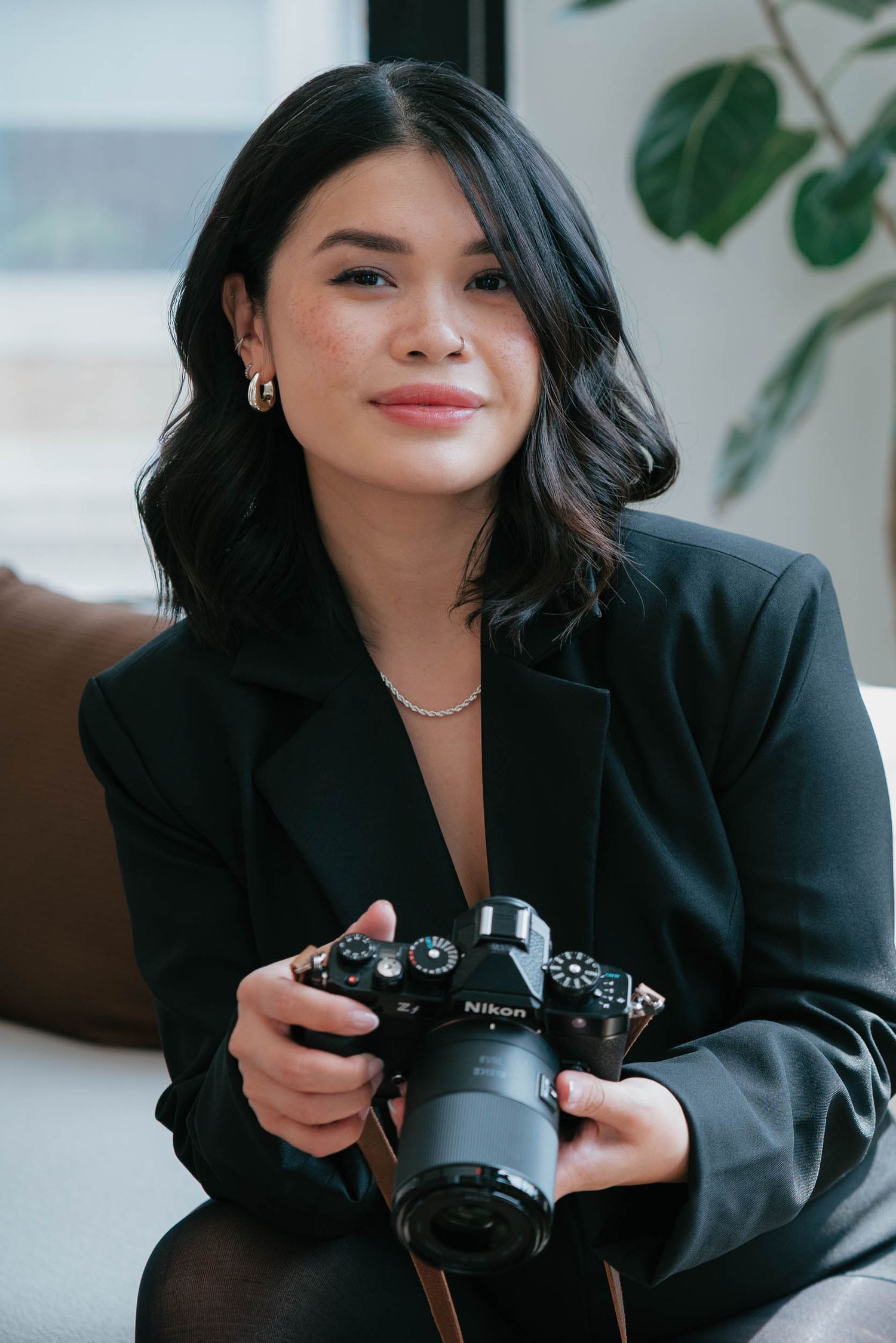 Young woman in black blazer holding a Nikon camera, smiling softly in a bright modern interior setting.