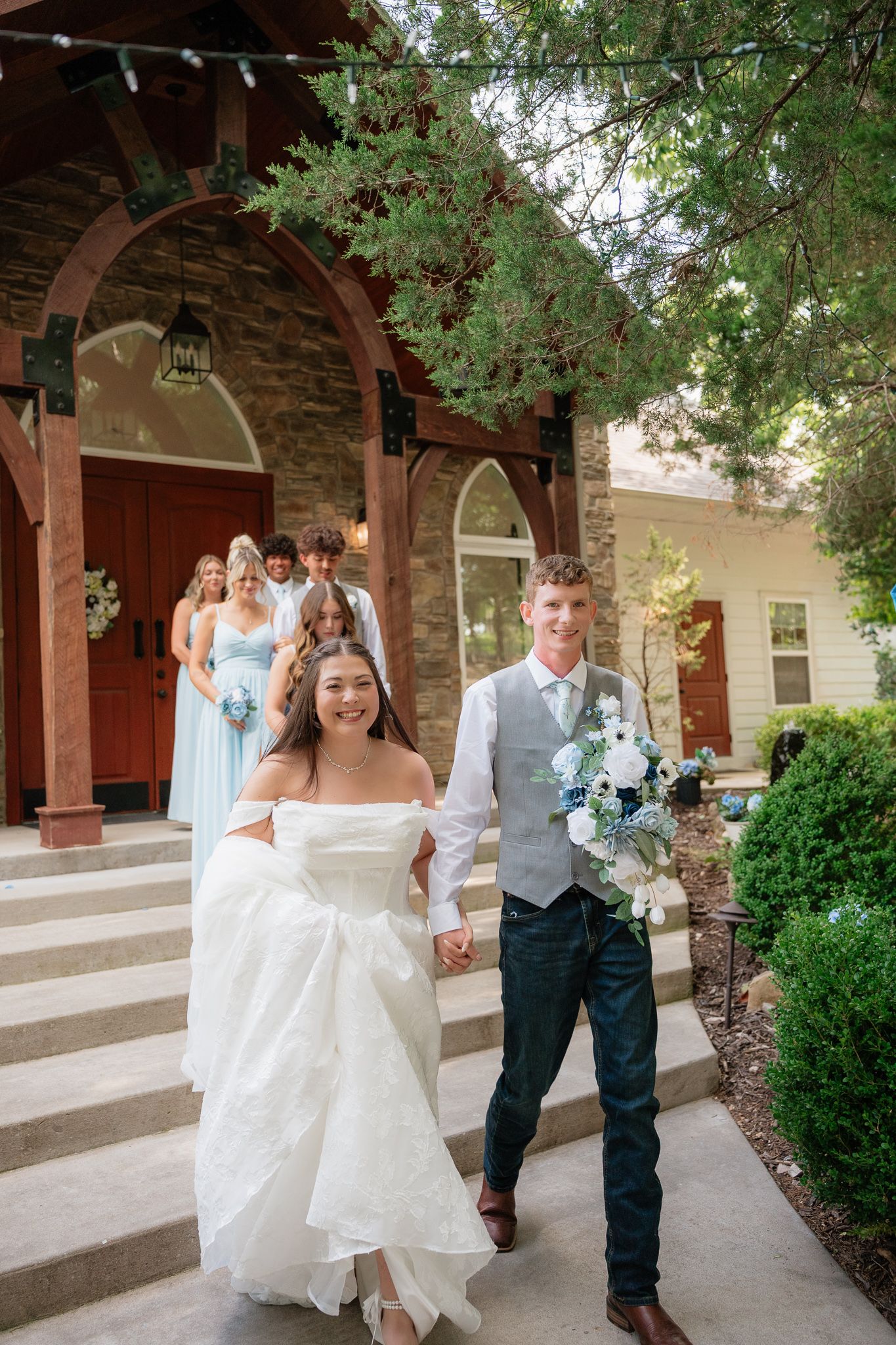 A smiling bride in a white gown and groom in a gray vest hold hands walking from a chapel with wedding party behind them.