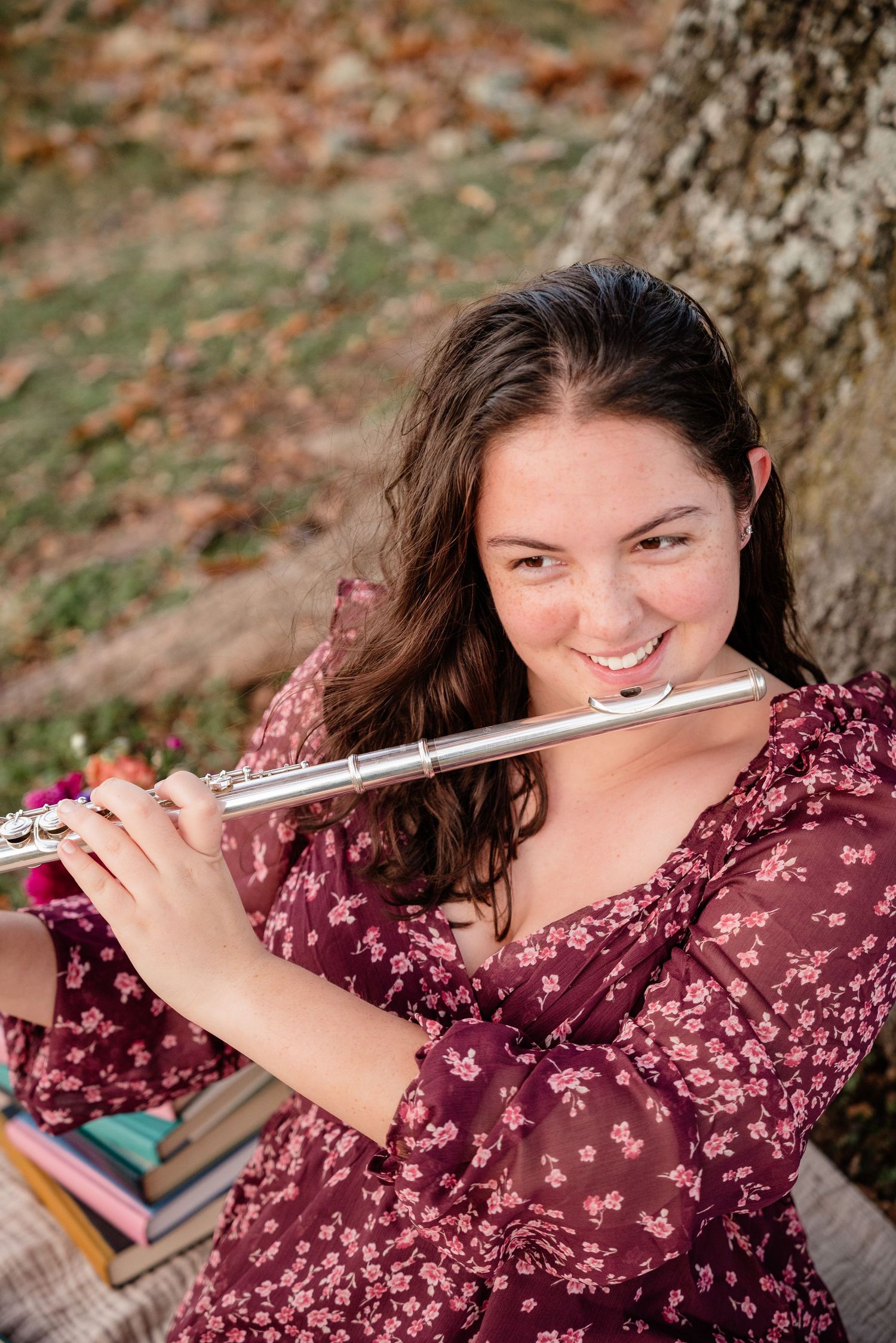 Young woman in floral dress playing silver flute outdoors near tree, smiling warmly at camera.