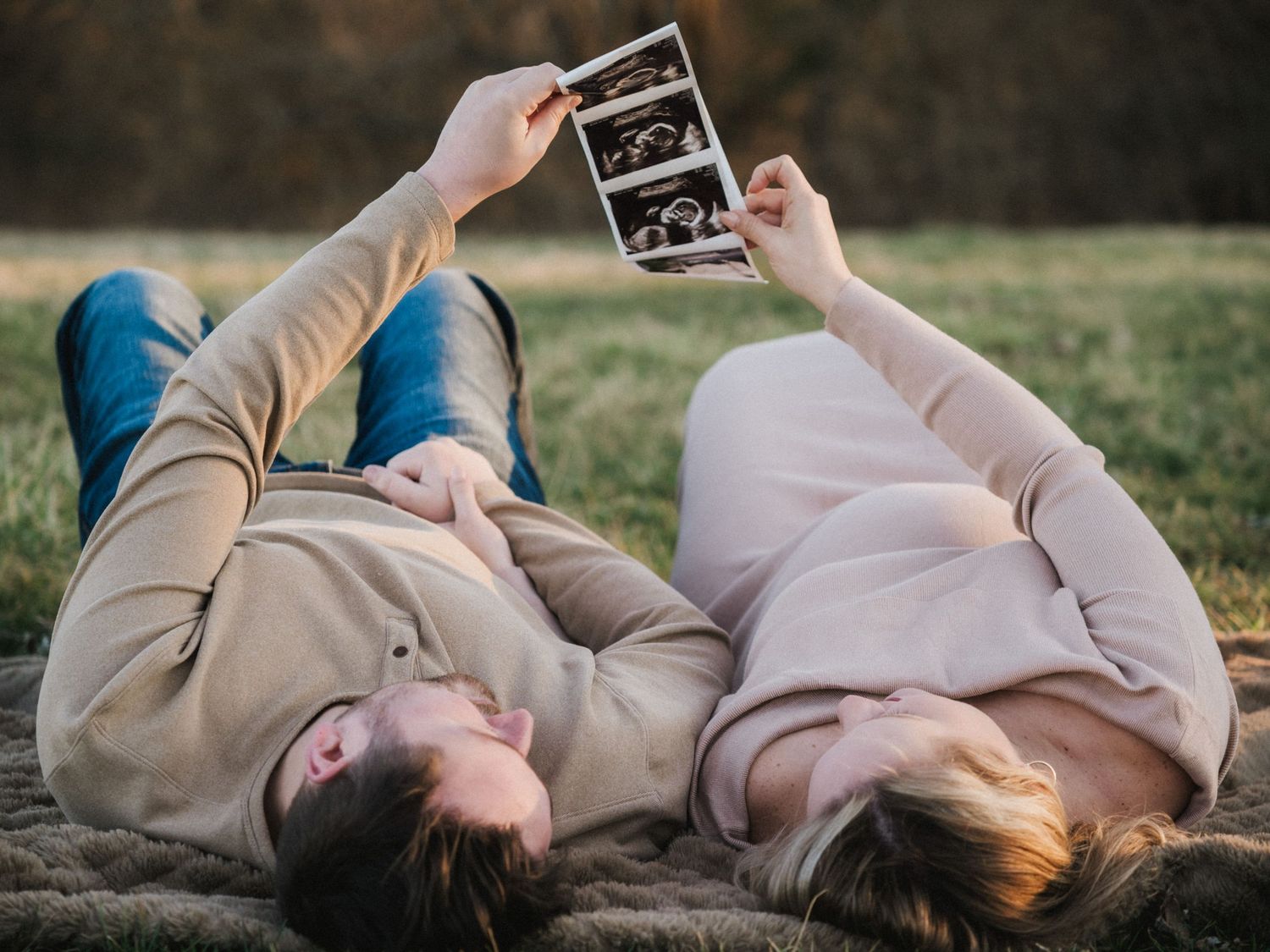 Couple lying on grass holding ultrasound photos, woman with baby bump, sharing pregnancy announcement moment outdoors.
