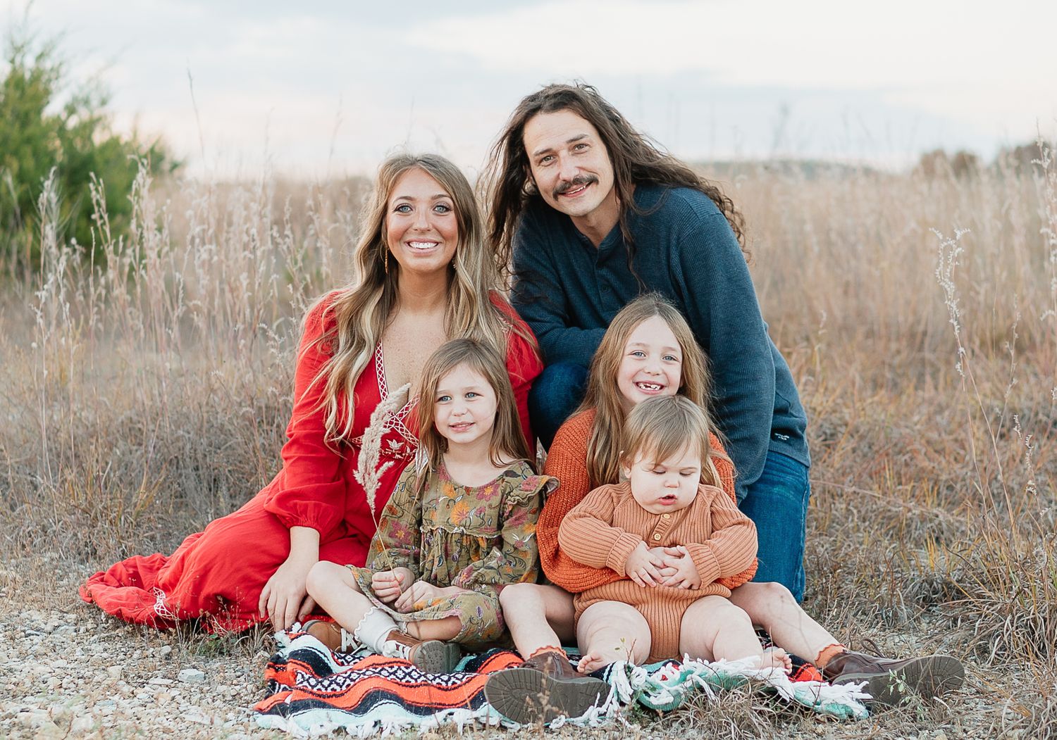 Family of five smiling outdoors in a field, parents and three young children sitting on a colorful blanket in nature.