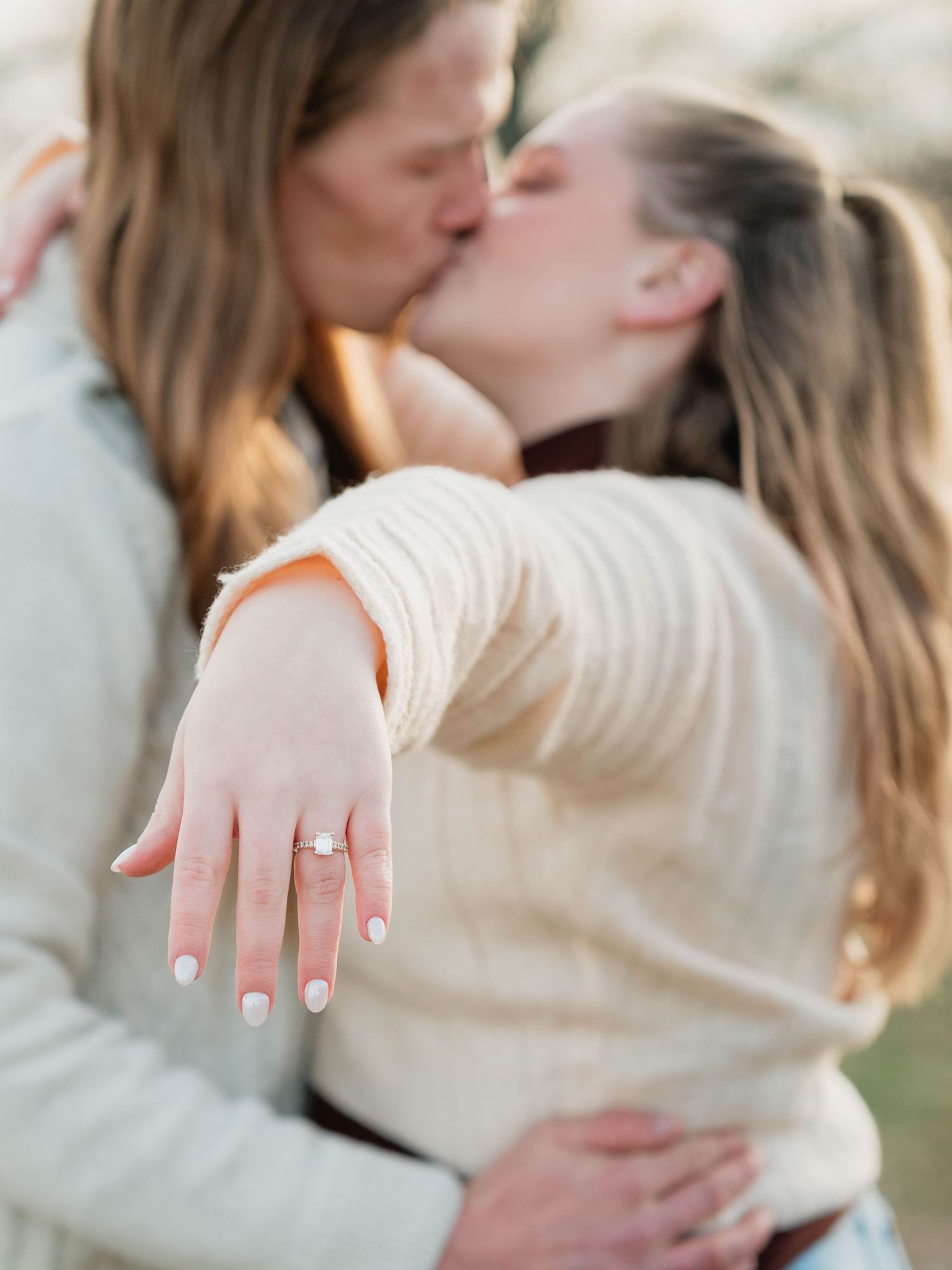 Couple kissing while showing off engagement ring with white French manicure during outdoor proposal photo session.