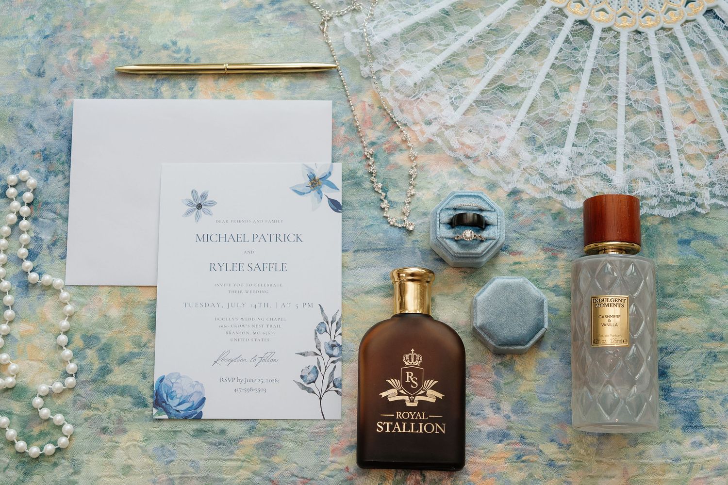 Wedding flat lay with floral invitation, Royal Stallion cologne, crystal perfume bottle, pearl necklace, and ring box on blue backdrop.