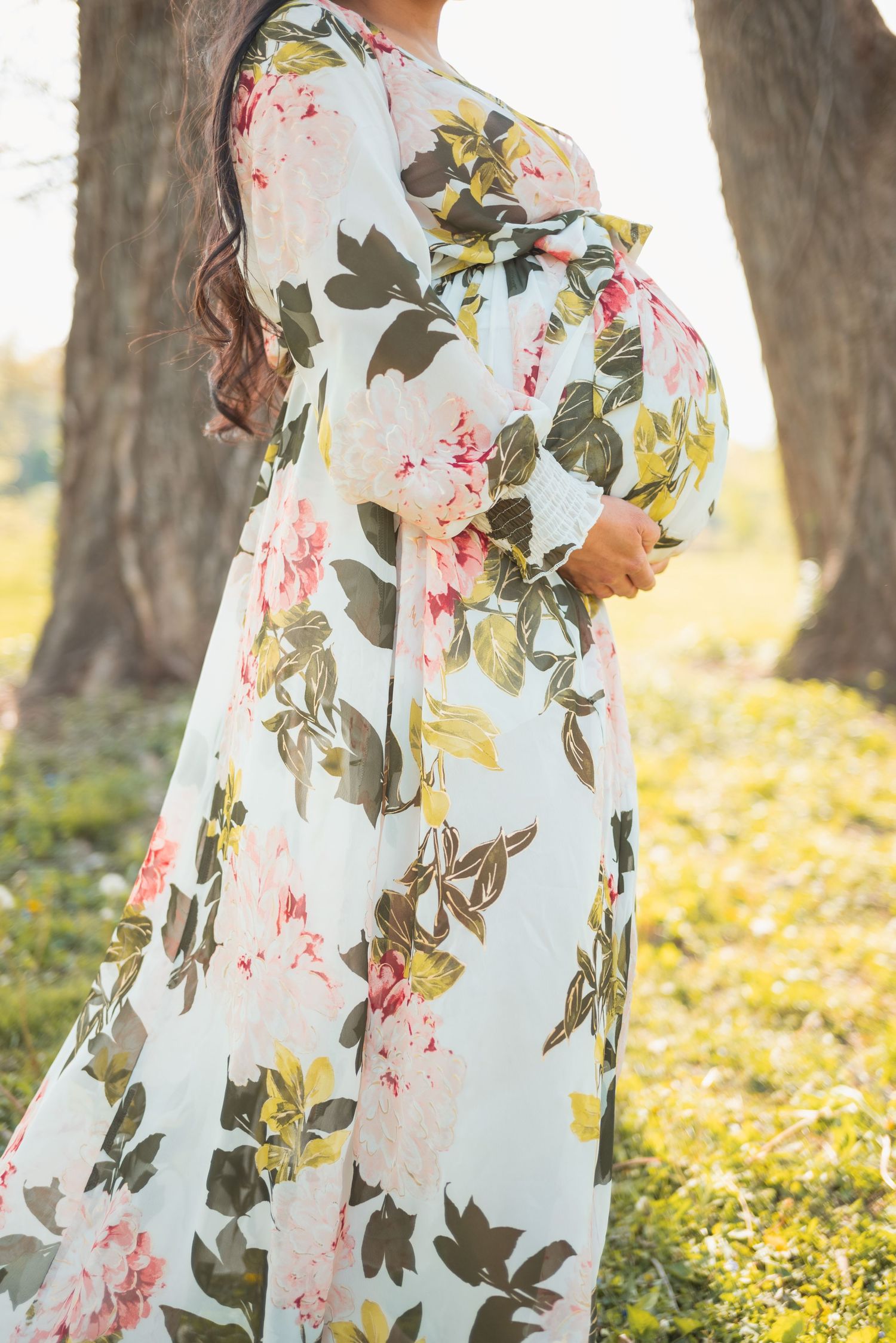Pregnant woman in floral maxi dress cradling baby bump outdoors in sunlit park setting.