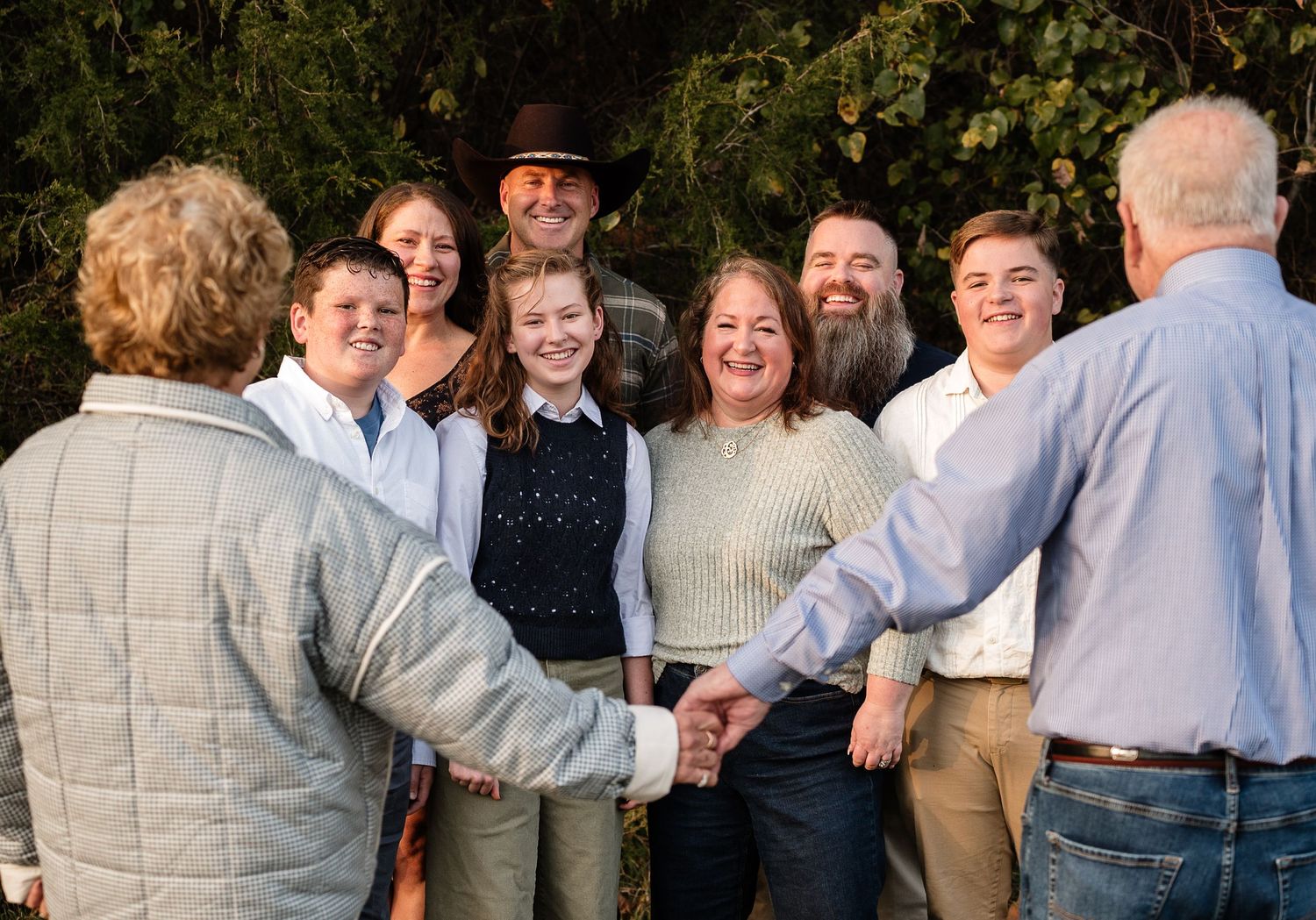 A joyful family group photo outdoors, smiling and shaking hands among lush green trees at dusk.