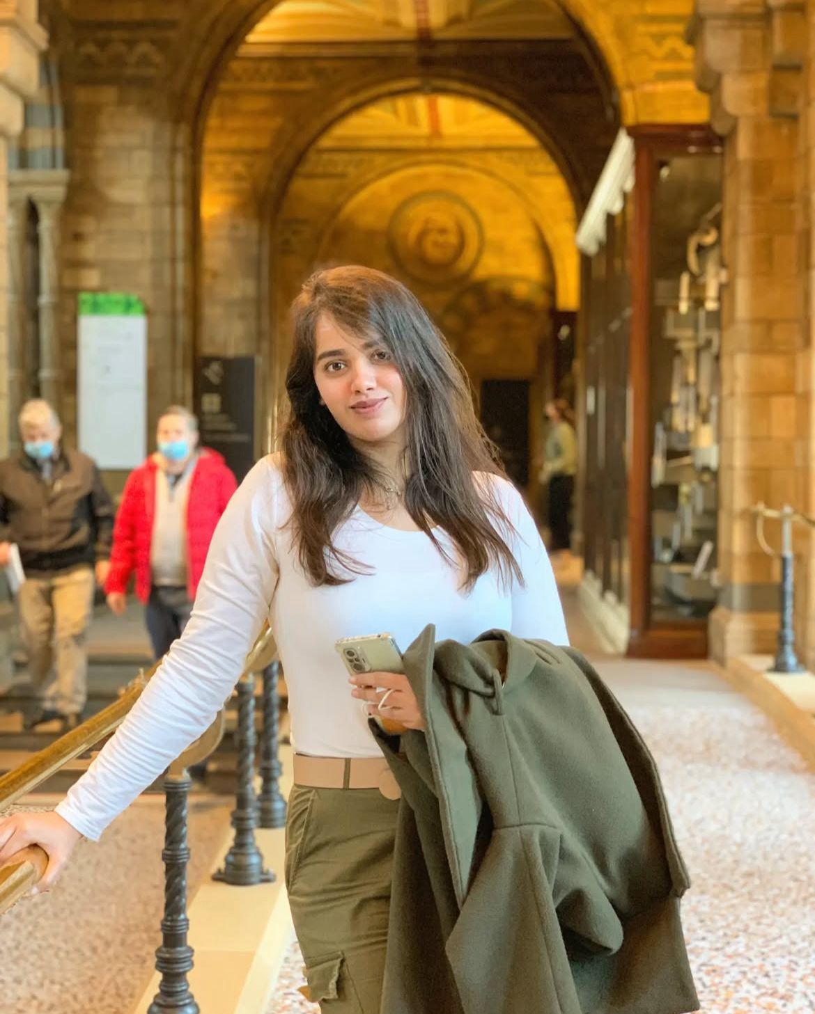 Young woman in white top and olive cargo pants holding a jacket inside a grand historic building in london.