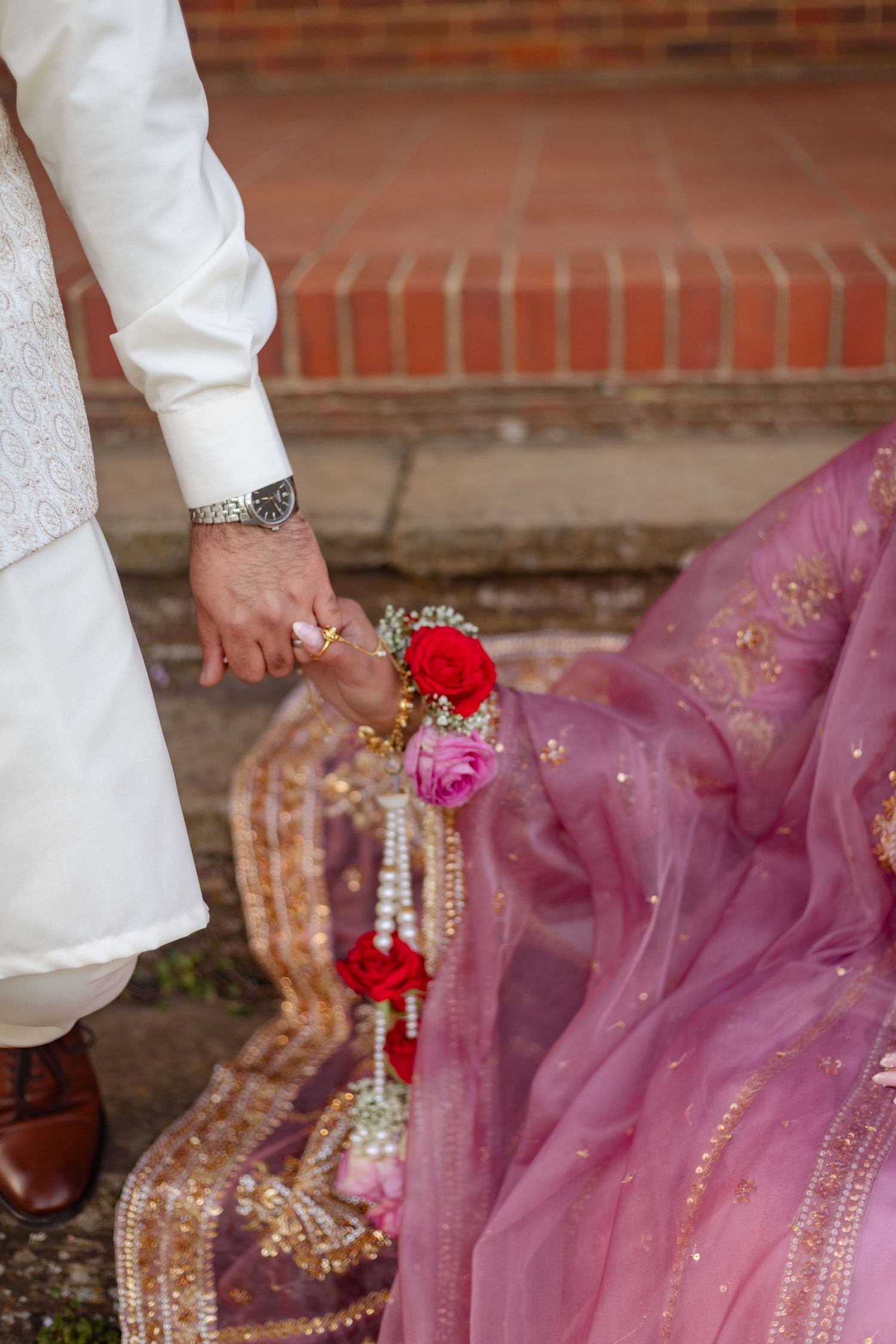 Groom in white sherwani holds floral garland beside bride in pink embroidered lehenga outdoors in london.