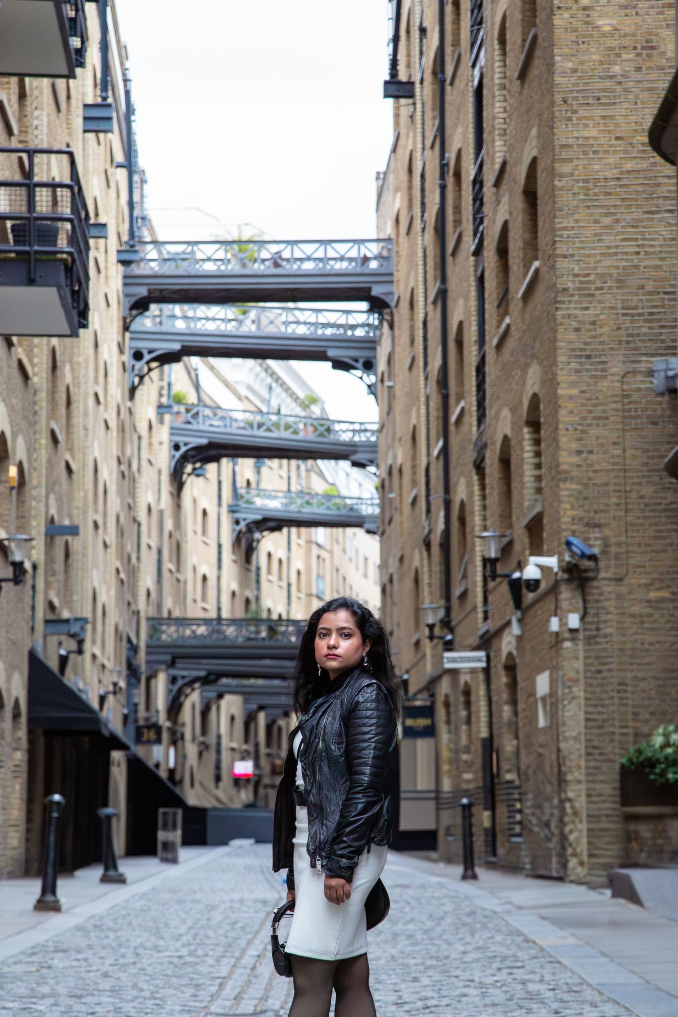 Young woman in leather jacket poses on cobblestone street between brick warehouses with iron bridges overhead in London.