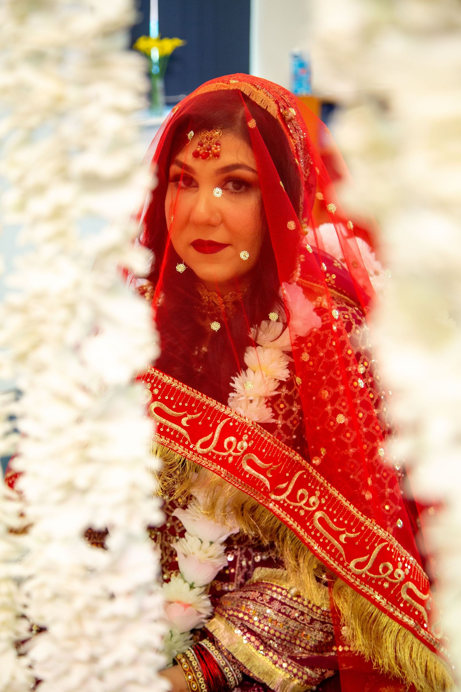 Beautiful South Asian bride in red bridal attire with gold jewelry and traditional veil indoors in London.