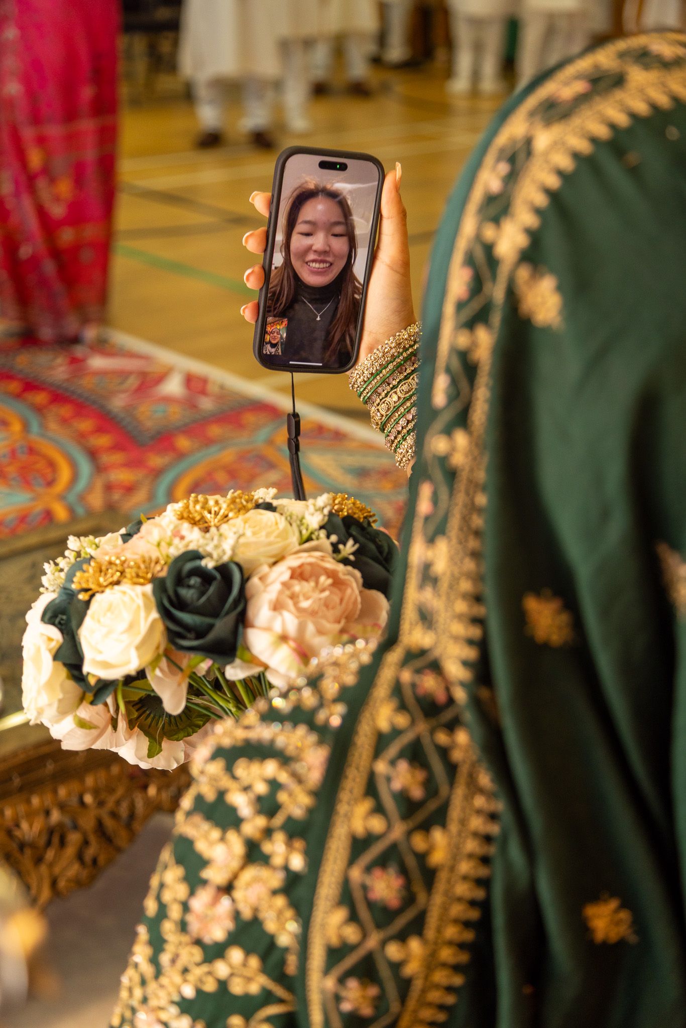 A bride in green embroidered attire holds flowers and FaceTimes a smiling woman during a traditional wedding ceremony.