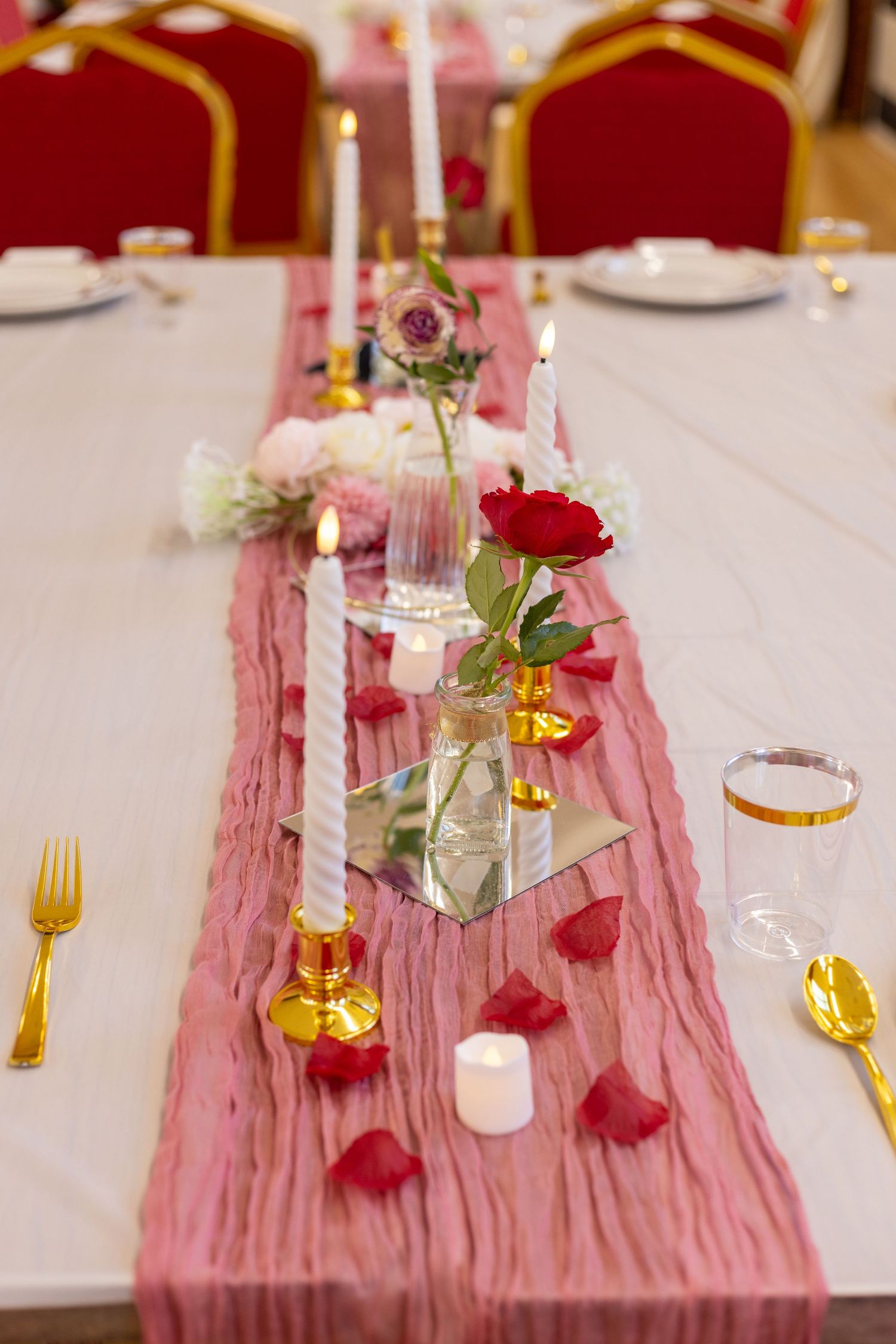 Elegant wedding table setting with pink runner, gold cutlery, red roses, candles, and rose petals on white tablecloth in london.