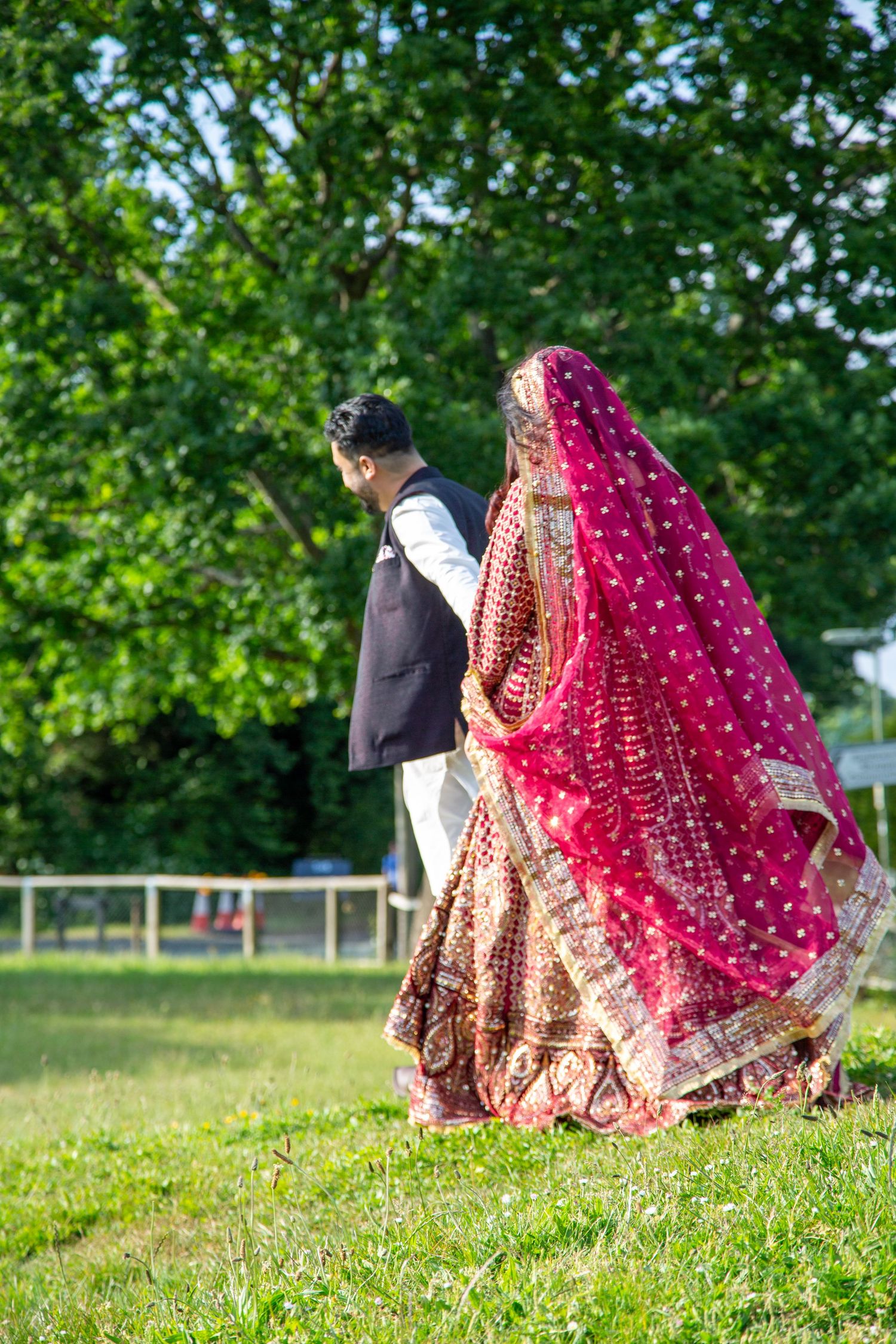 Pakistani bride and groom walking outdoors, bride wearing stunning red embroidered lehenga with dupatta in London.
