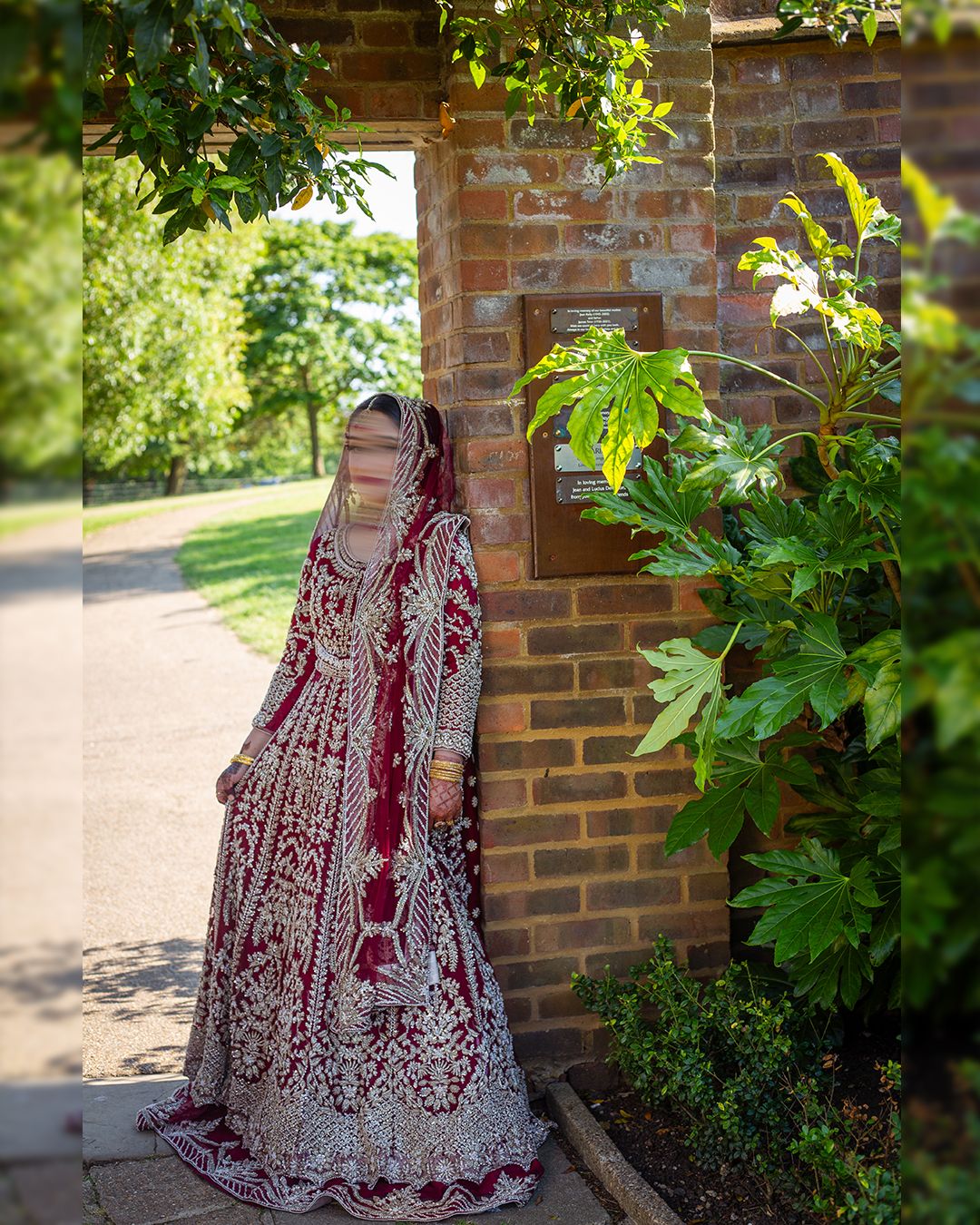 Pakistani Bride in ornate red and silver embroidered lehenga with dupatta stands beside brick wall and lush green plants in north london.