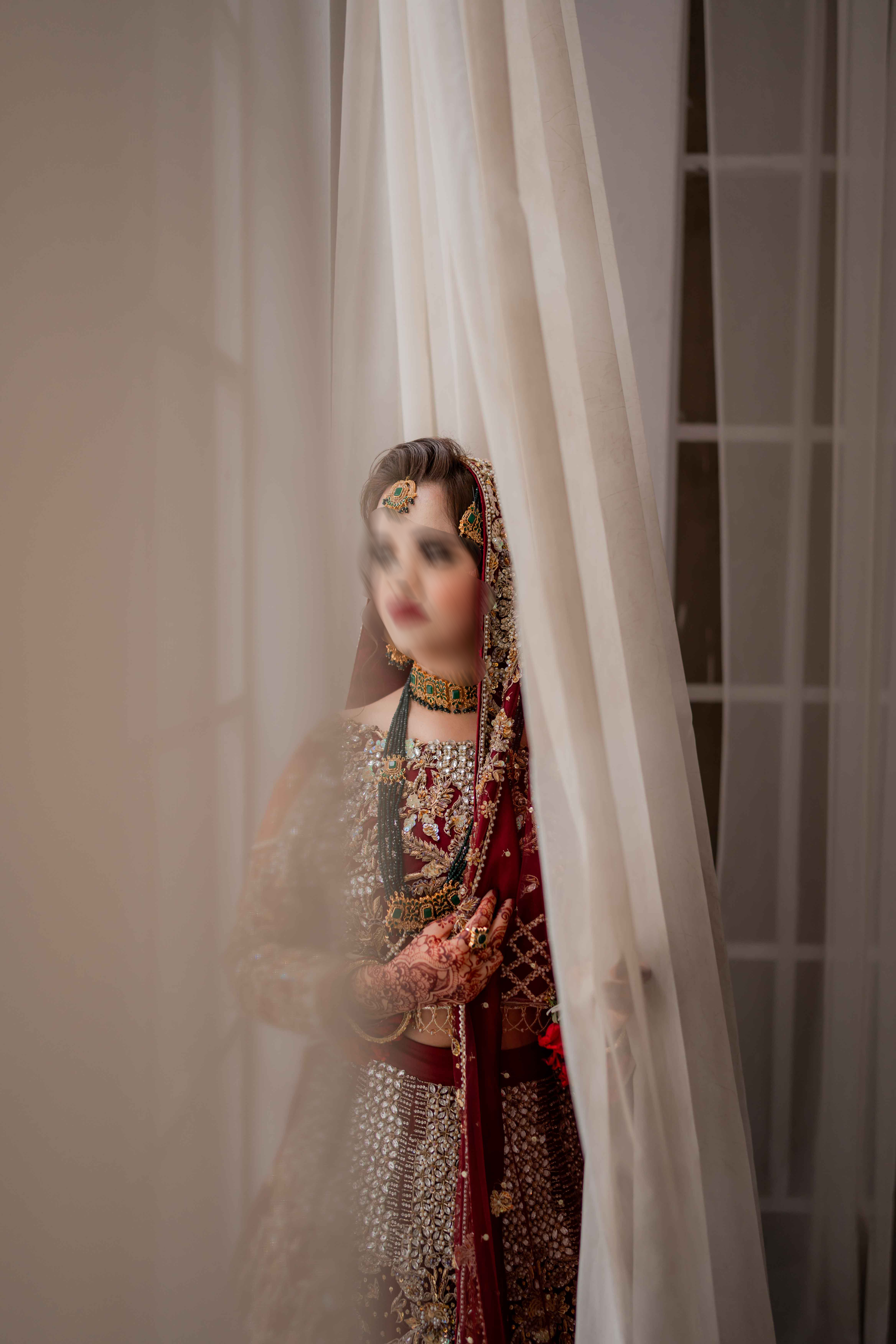 South Asian bride in red bridal lehenga with gold jewelry peers through sheer white curtains in london.