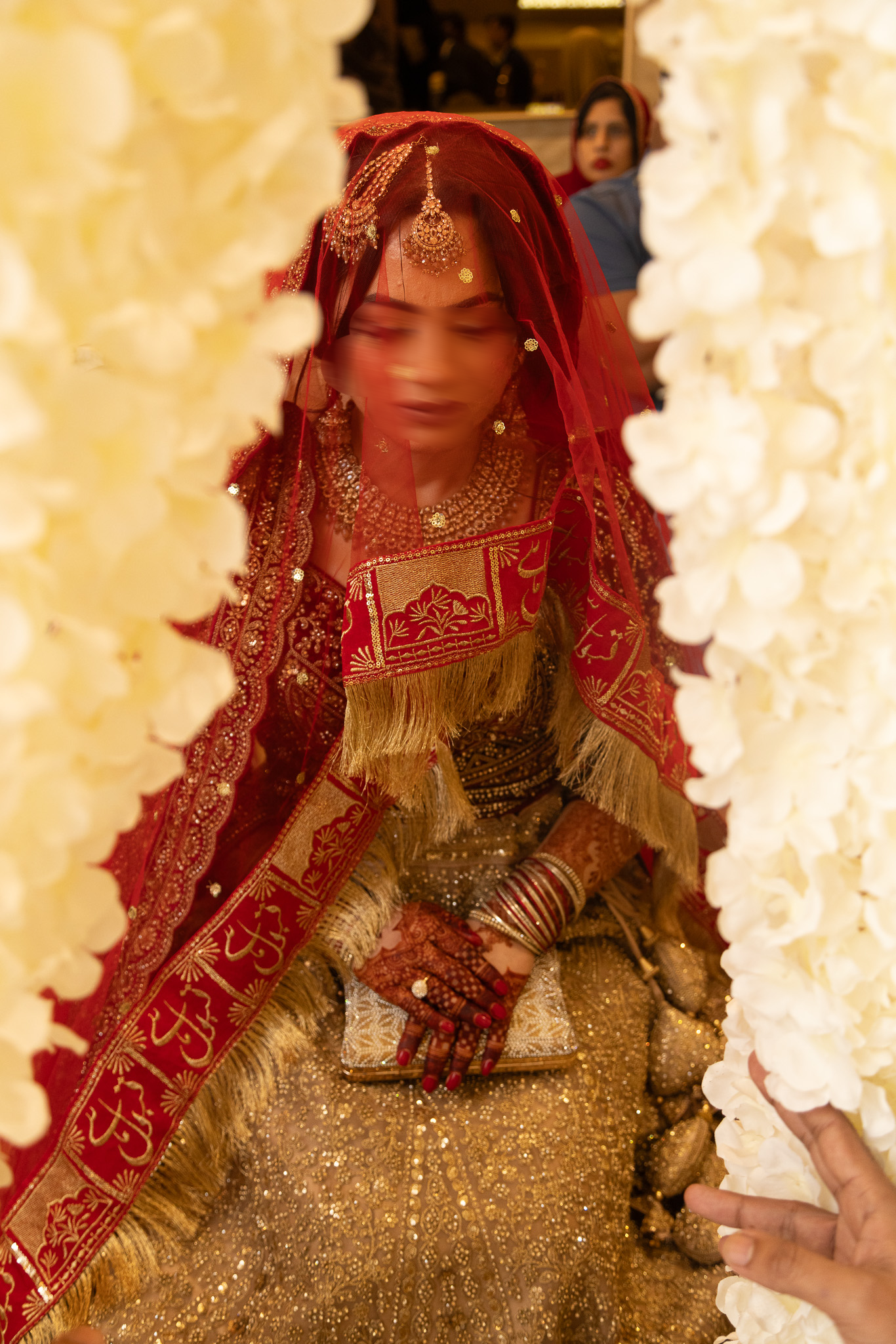Pakistani bride in gold and red bridal lehenga walking through white floral arch at wedding in london.