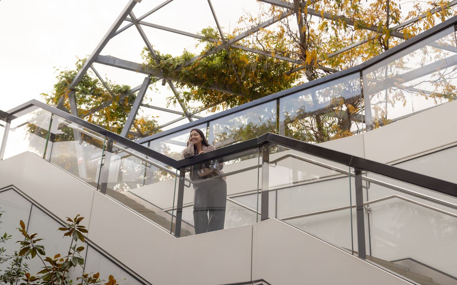 Woman standing on modern glass balcony staircase surrounded by lush green foliage overhead in london