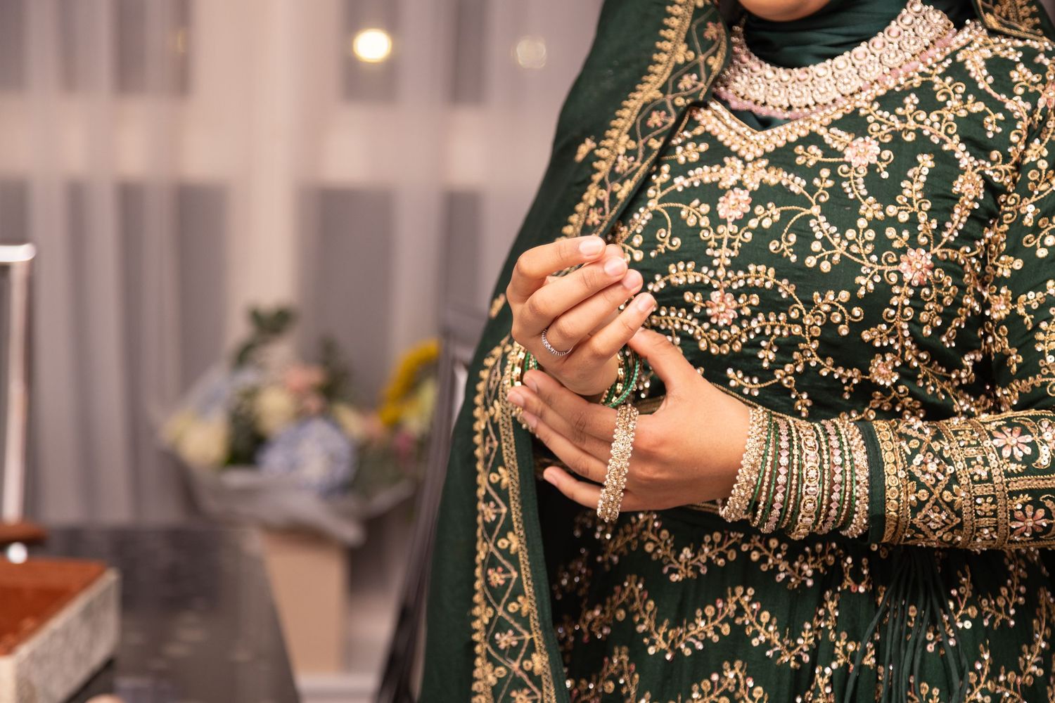 Woman in embroidered green Bangali bridal outfit with gold bangles and mehndi, hands clasped in prayer pose in London
